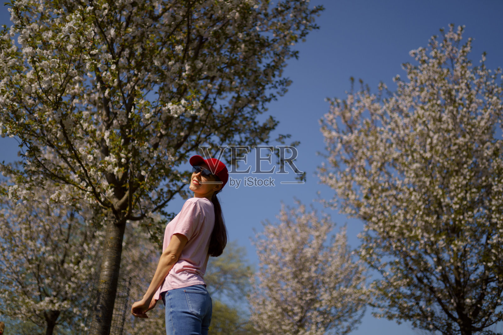 成功的商业女性穿着牛仔裤、粉色t恤和一顶时尚的红色帽子，在樱花盛开的公园享受她的休闲时间照片摄影图片