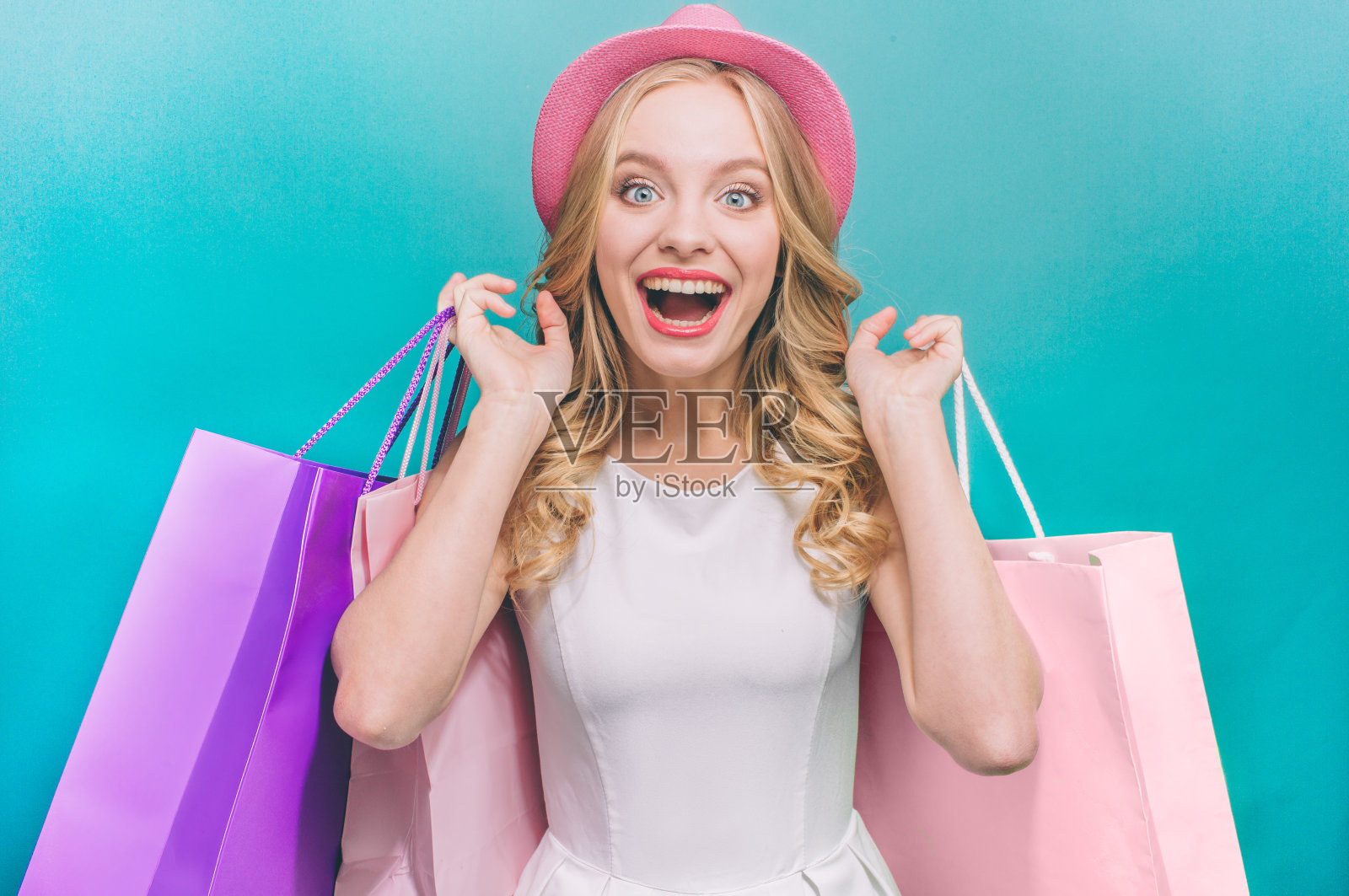 开朗迷人的女孩背着五颜六色的包包。她还戴着粉红色的帽子。孤立在蓝色背景。照片摄影图片