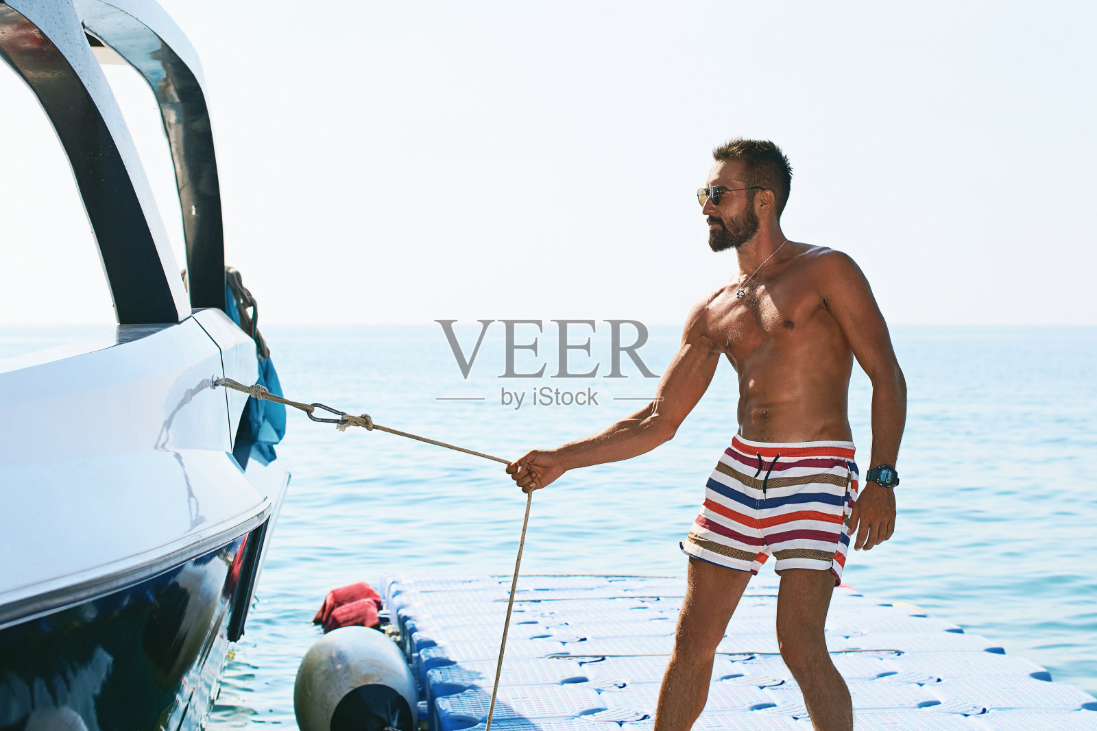 年轻帅气的穆斯林船长穿着短裤，拉着游艇或船的绳索，站在塑料浮桥上照片摄影图片