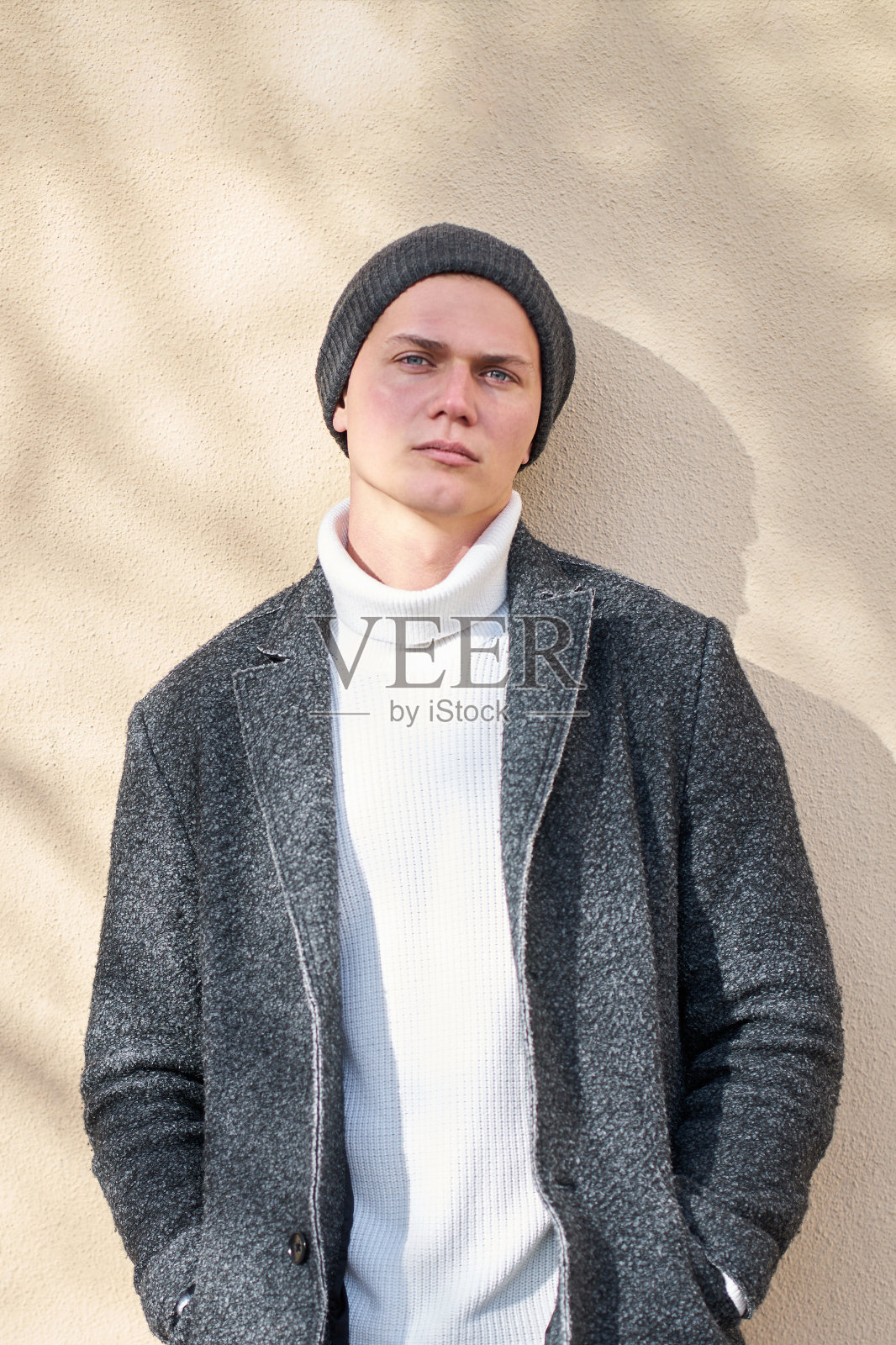 现代都市年轻严肃时髦的男人穿着现代灰色外套，白色时髦毛衣和黑色牛仔裤站在米色墙壁背景和看着相机。照片摄影图片