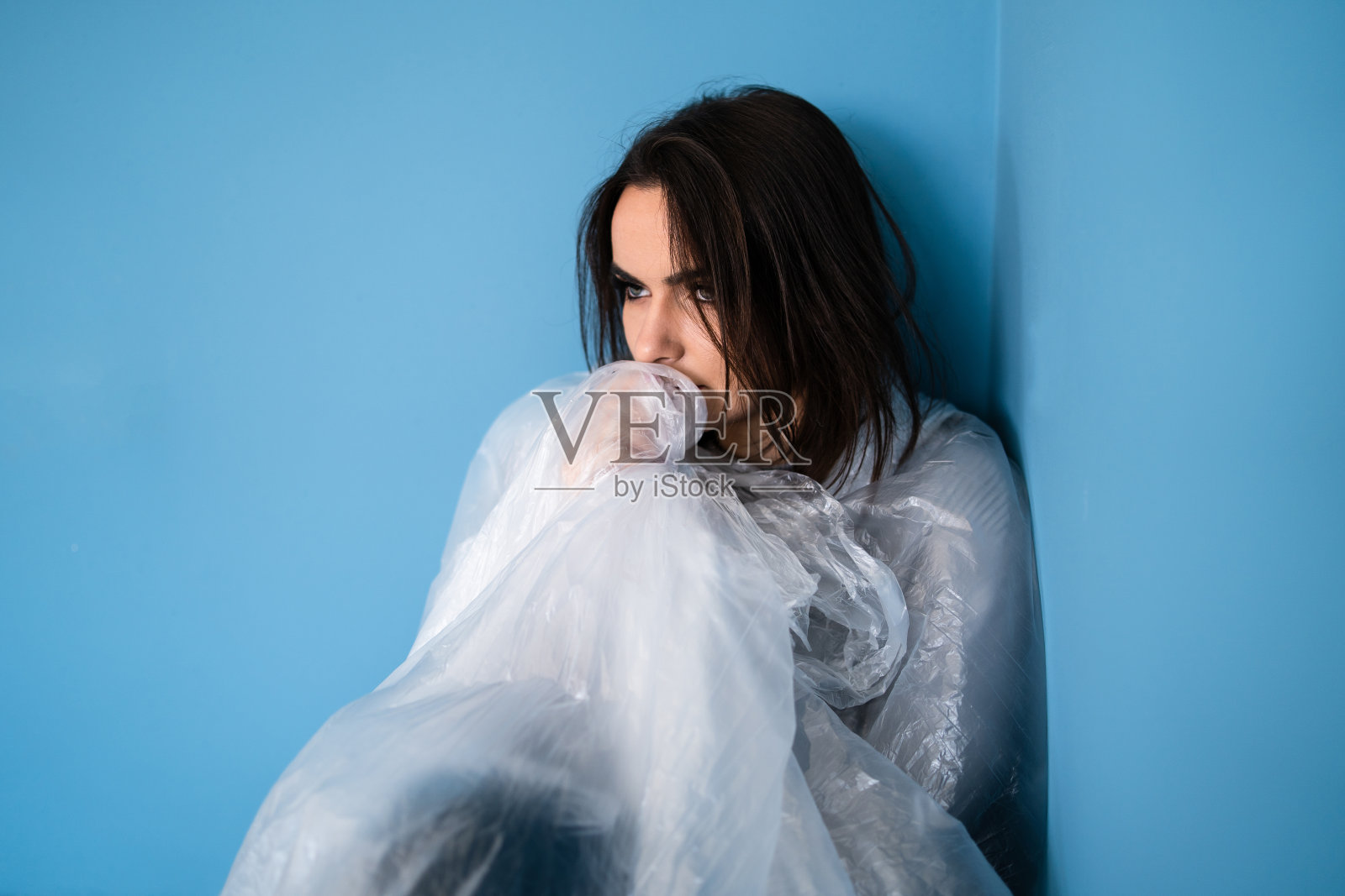 年轻的害怕的女人悲伤的覆盖着塑料尼龙玻璃纸无助地坐在一个角落的蓝色房间污染拯救地球的概念照片摄影图片