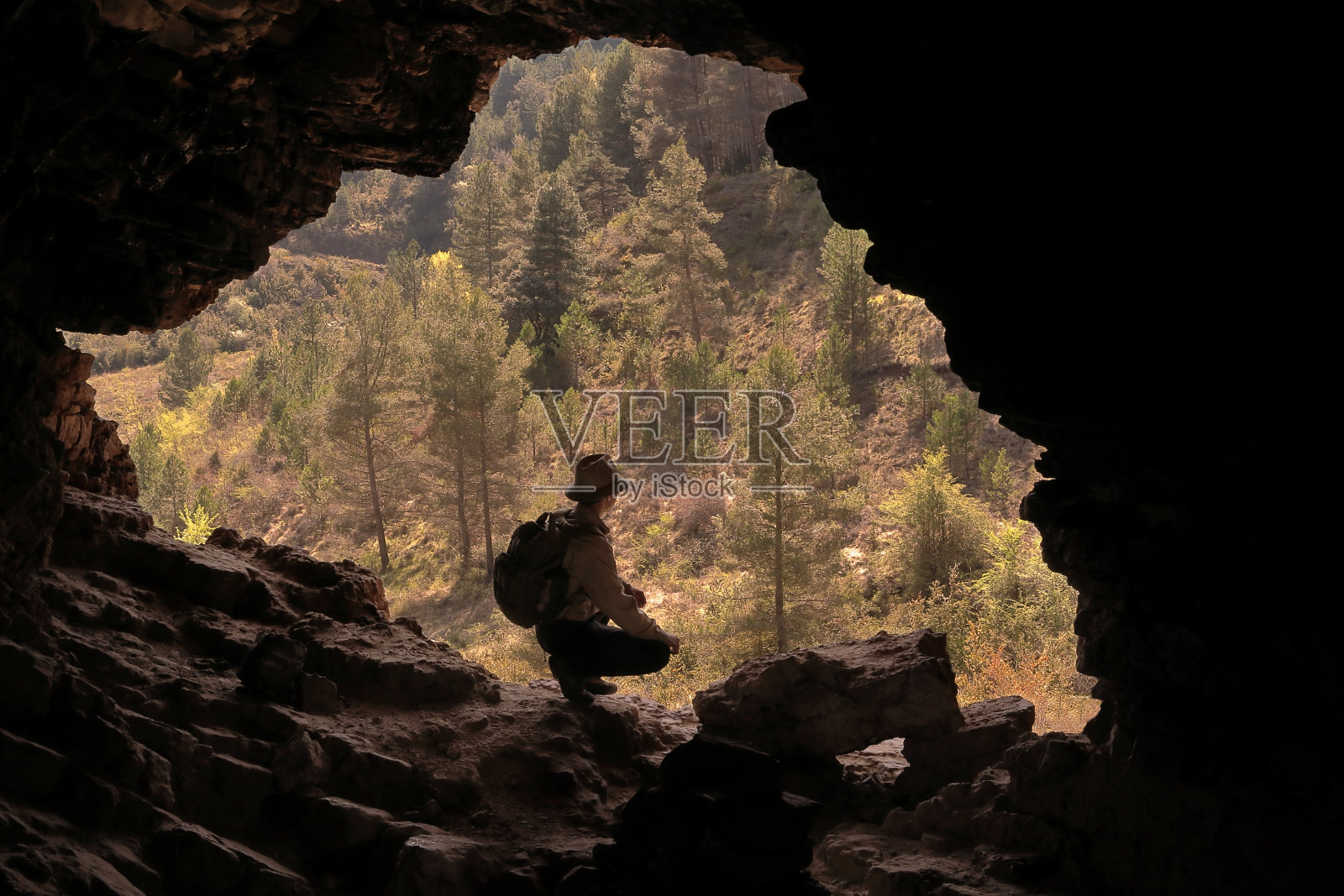 戴着帽子和背包的徒步旅行者和冒险家在一个山洞里休息，看一个绿树环绕的美丽山谷照片摄影图片