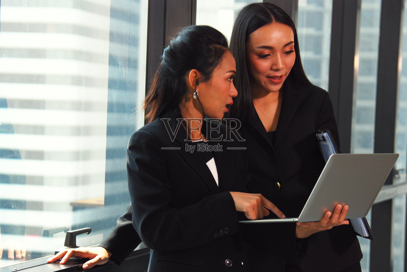 两个漂亮的女商人在办公室一边聊天一边玩笔记本电脑。照片摄影图片