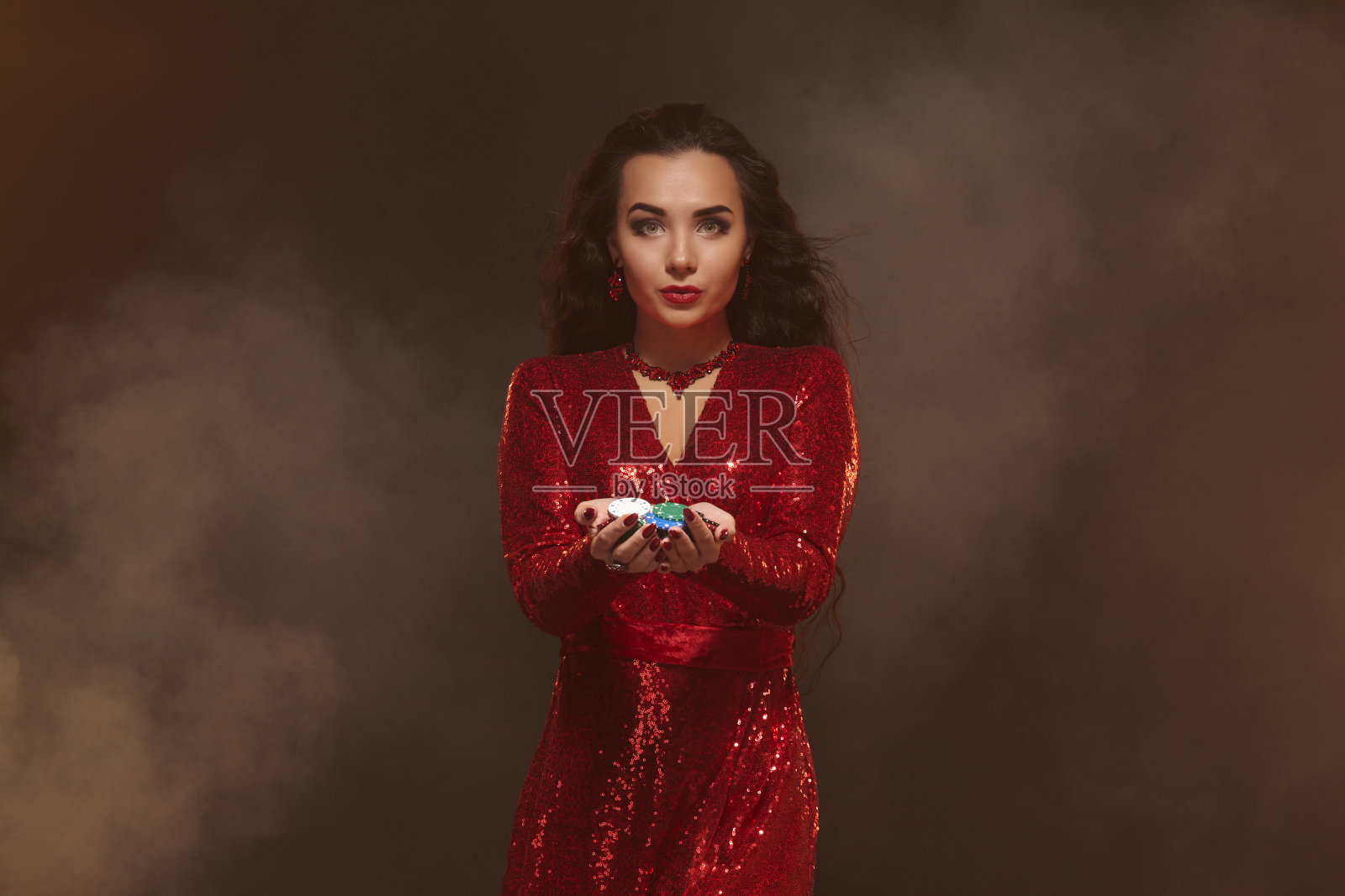 年轻漂亮幸运的黑发女人穿着红色晚礼服，手里拿着扑克筹码，显示她的奖金。照片摄影图片