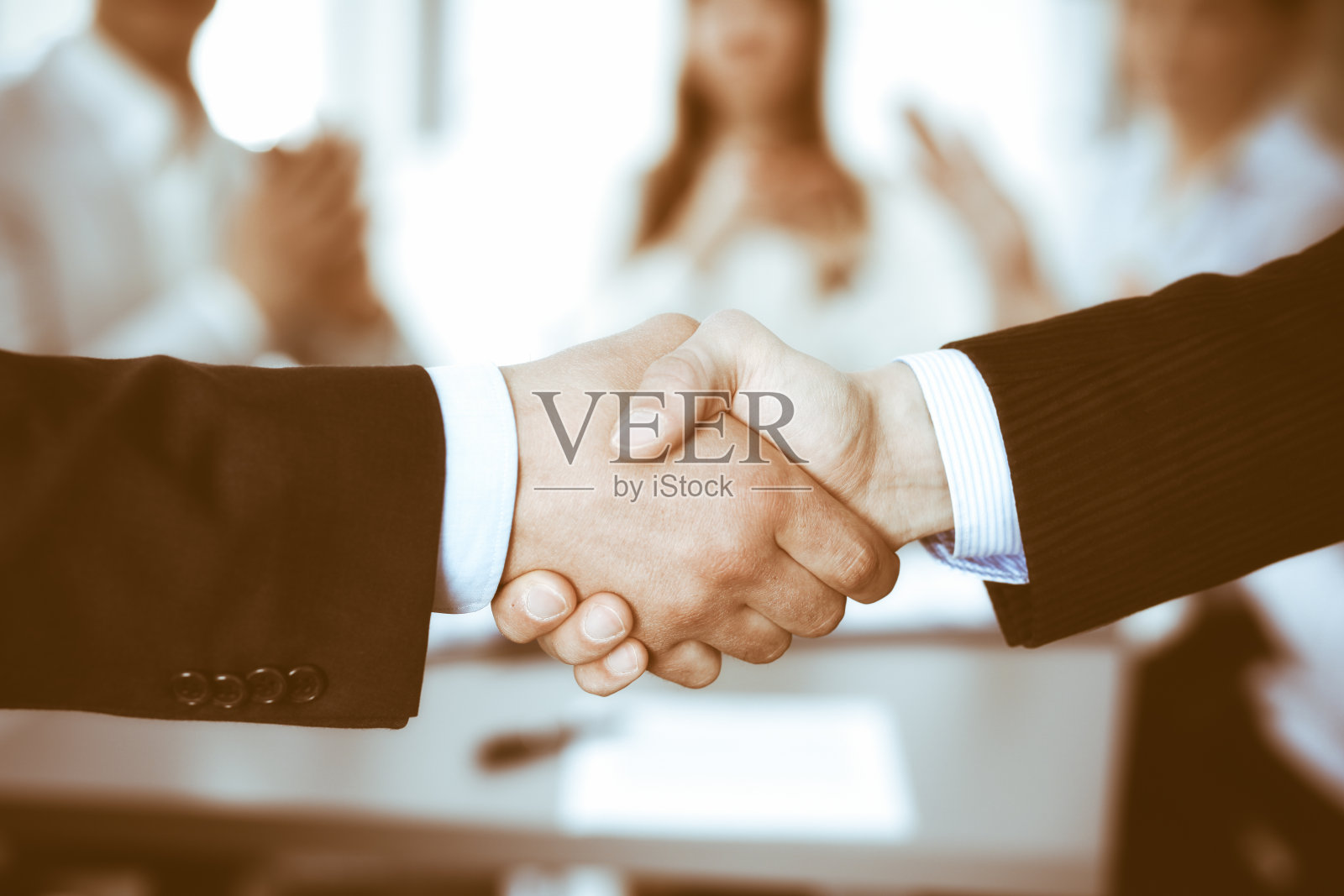 商务人士在会议上握手，而他们的同事在鼓掌和鼓掌。一群不知名的商人和妇女在现代白人办公室。成功的团队合作、伙伴关系和握手理念照片摄影图片