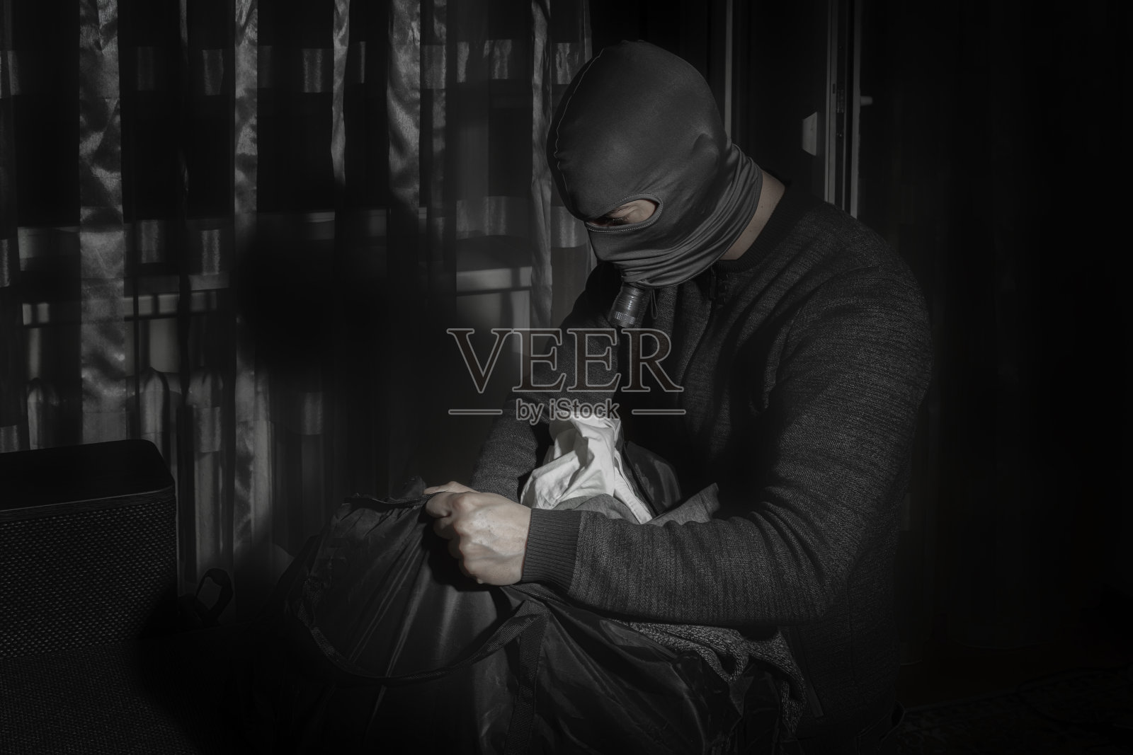 小偷戴着面具，拿着手电筒在房间里把赃物放在一个大袋子里照片摄影图片