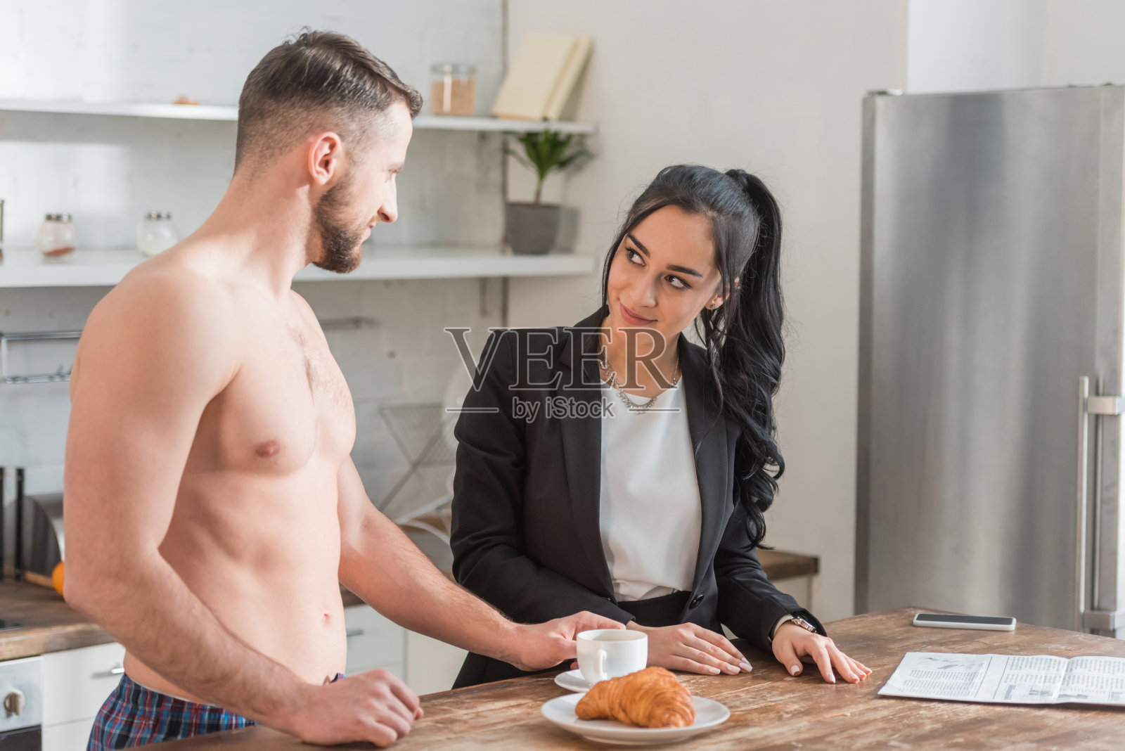 英俊的赤膊男子站在茶碟旁边，拿着牛角面包和一杯咖啡，看着迷人的女孩照片摄影图片