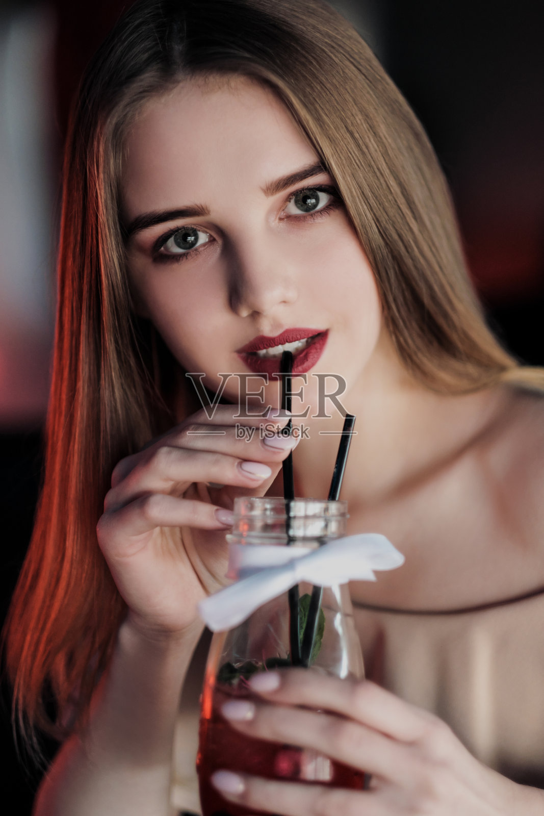 一个年轻漂亮的棕色头发的女孩坐在霓虹灯光下的咖啡馆里，坐在杯子旁边喝着红色的果汁。照片摄影图片