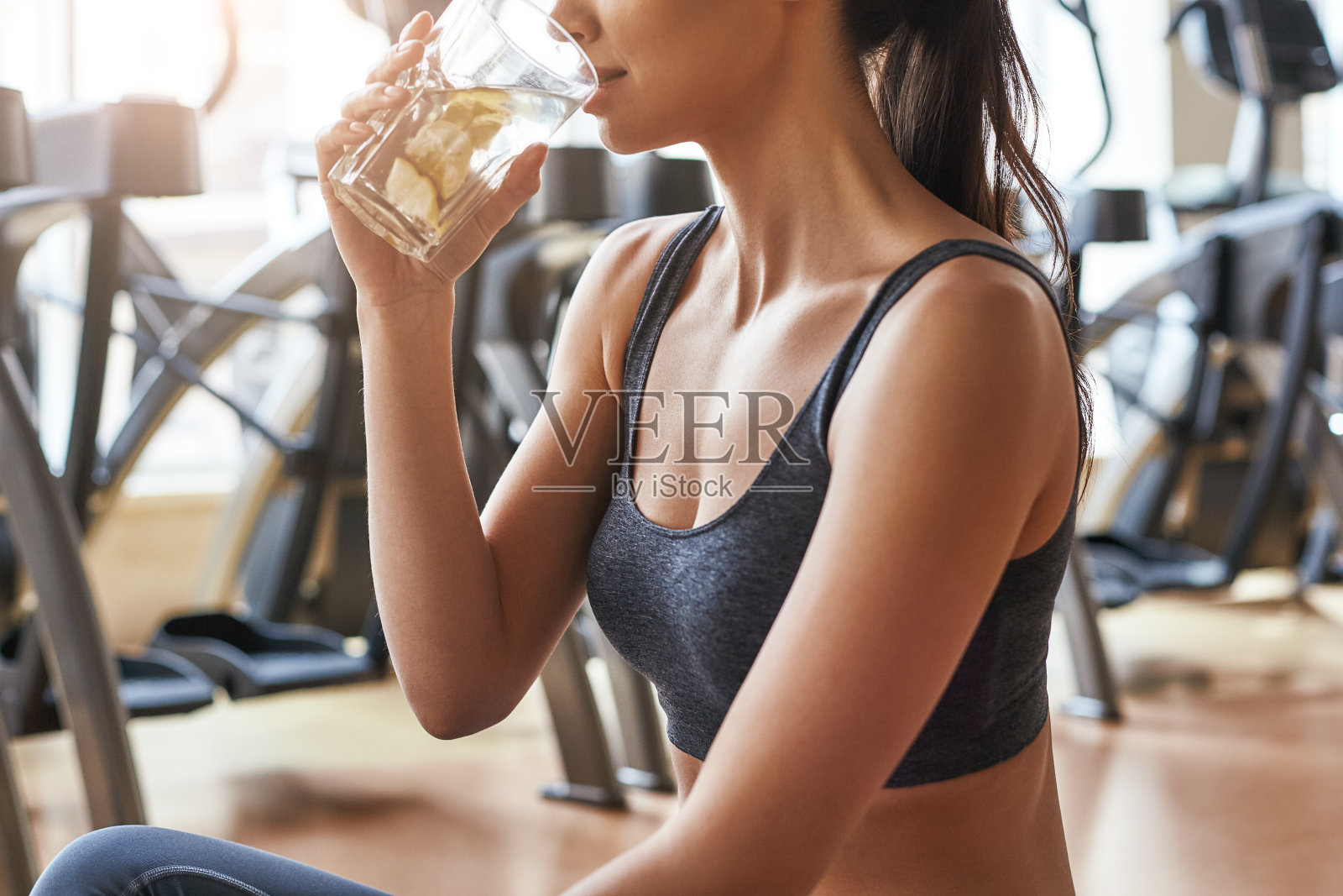 我需要水。裁剪的照片美丽和快乐的年轻女子在运动服装喝柠檬水，而坐在一个垫子在健身房照片摄影图片