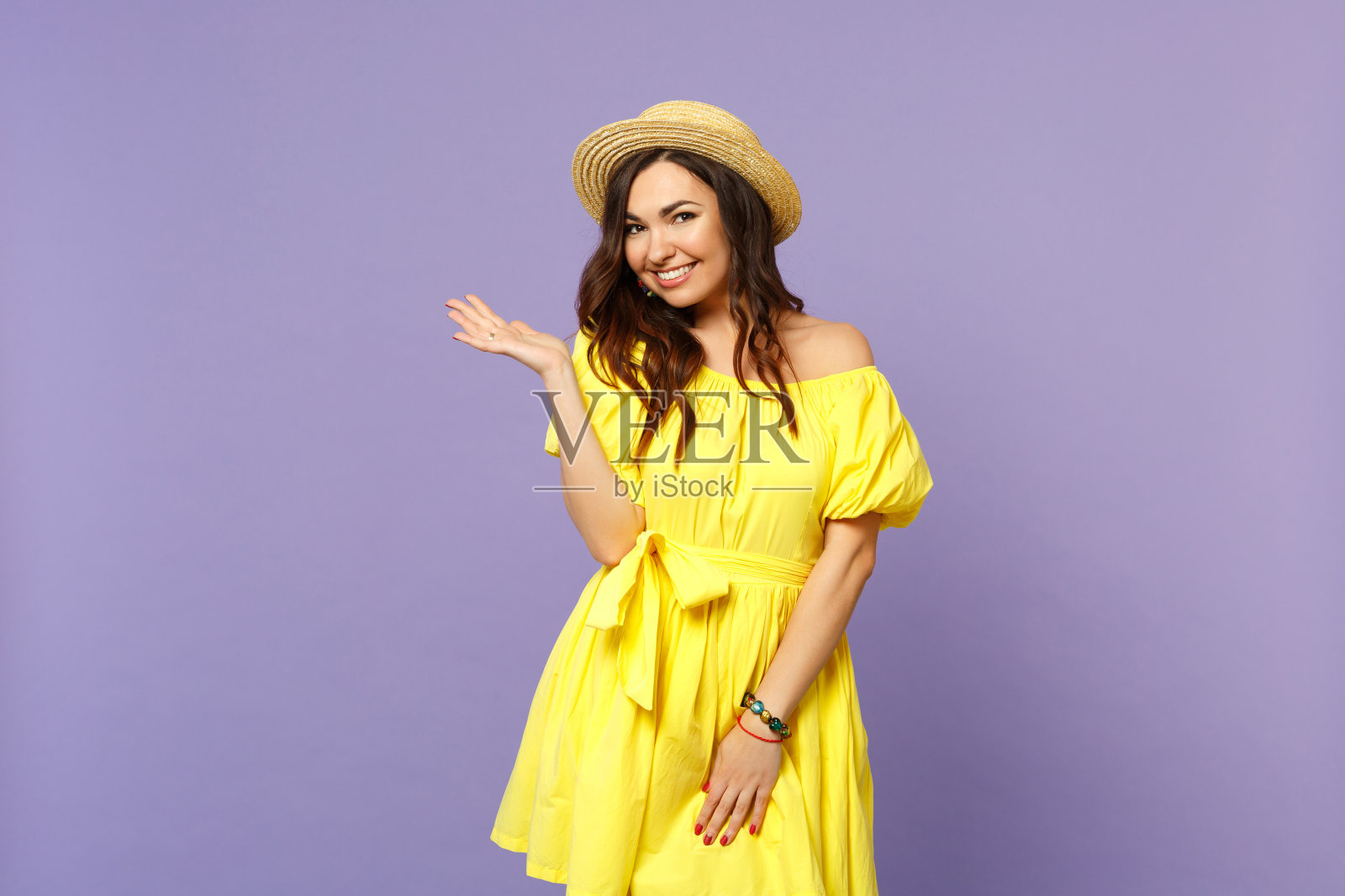 微笑的年轻女子的肖像在黄色的衣服，夏天的帽子看相机，指向一边孤立在柔和的紫罗兰背景。人真诚情感，生活理念。模拟拷贝空间。照片摄影图片