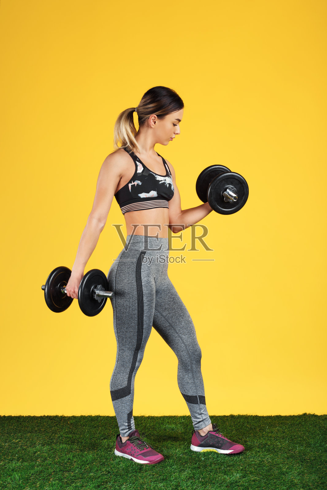 漂亮的健康的年轻女子与哑铃二头肌在绿色的草地在黄色的背景。照片摄影图片