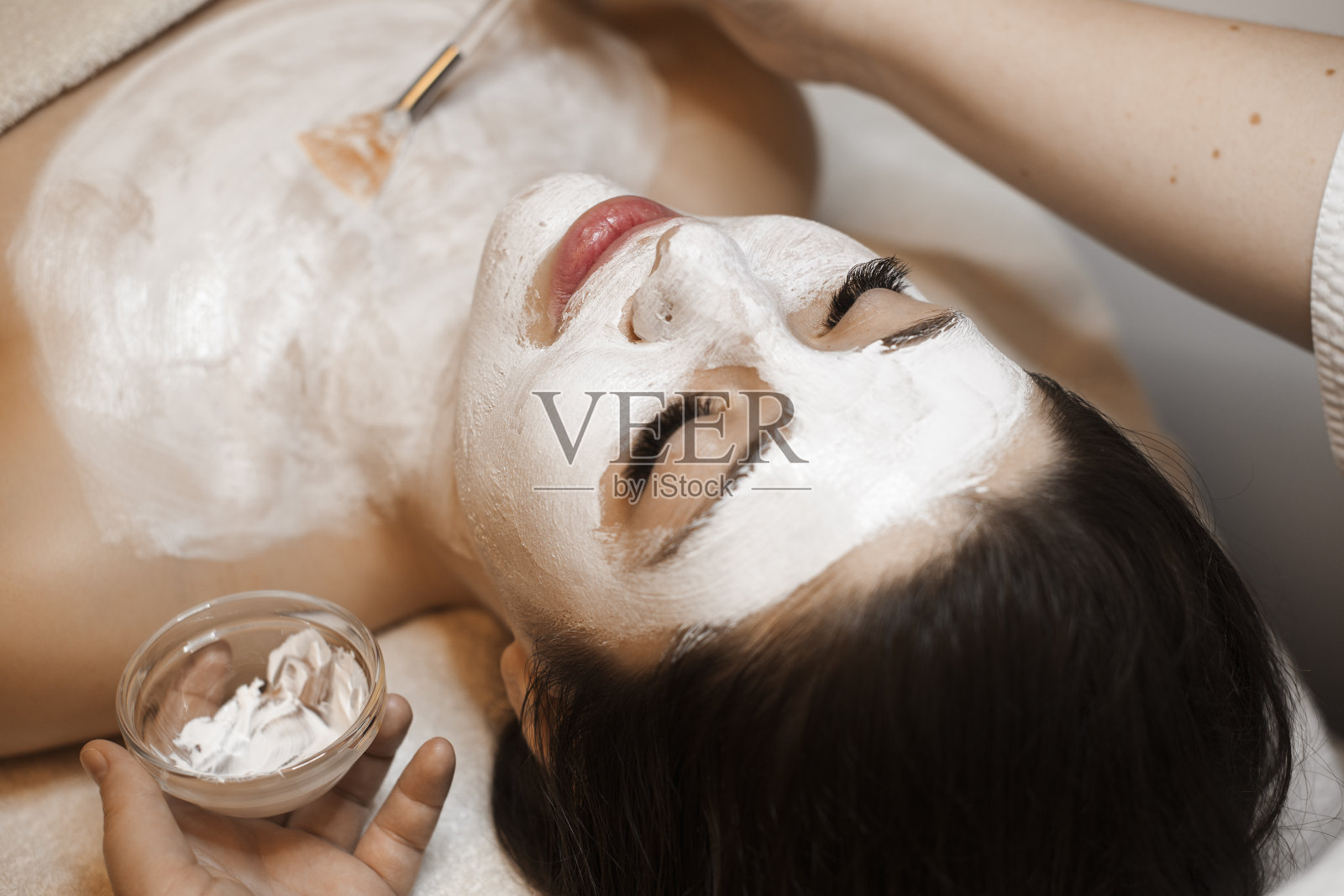 一个可爱的女人与黑色头发有一个白色面具在她的脸在一个健康水疗沙龙的上视图的近距离观察。照片摄影图片