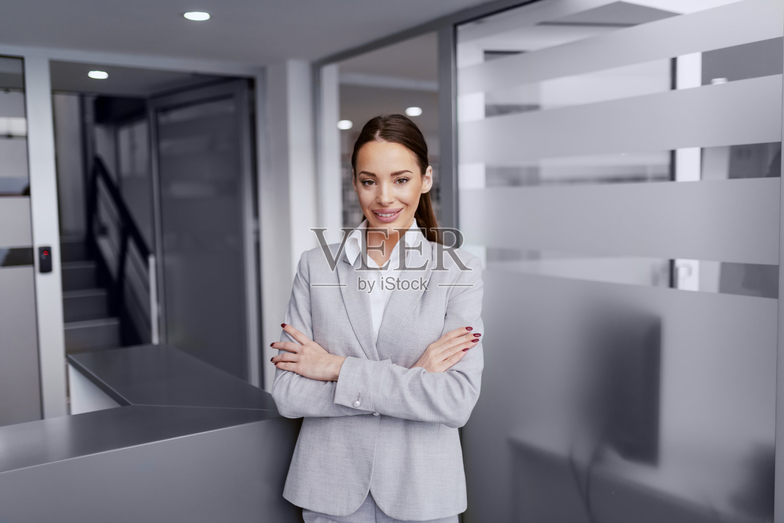 美丽的白人女商人与棕色长发和inn正式穿着站在大厅双臂交叉。灰色照片摄影图片