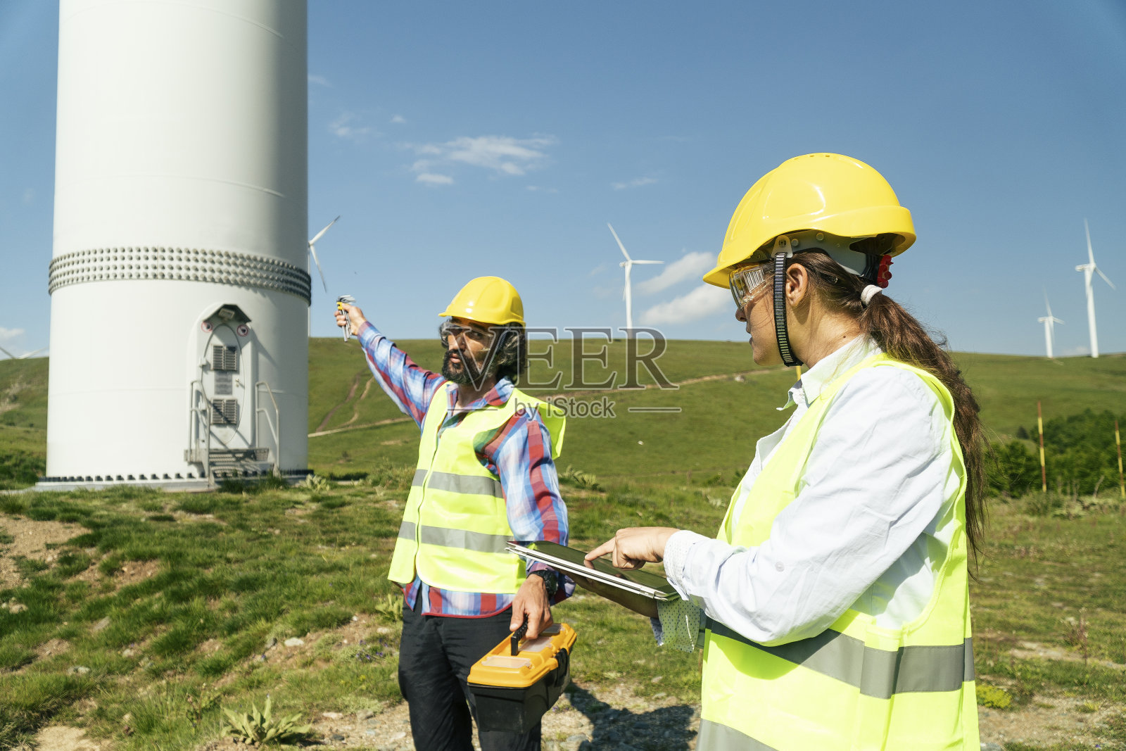 风力涡轮机农场的可再生能源系统工程。电力维护工程师在风力涡轮机发电站附近工作，身后是晴朗的蓝天。照片摄影图片
