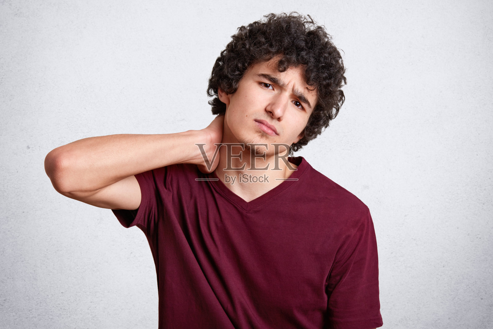 肖像的悲伤英俊的男人，小胡子和黑色的卷发，穿着休闲的栗色t恤，站着，握住他痛苦的脖子。工作室拍摄，模型摆姿势孤立在白色背景。照片摄影图片