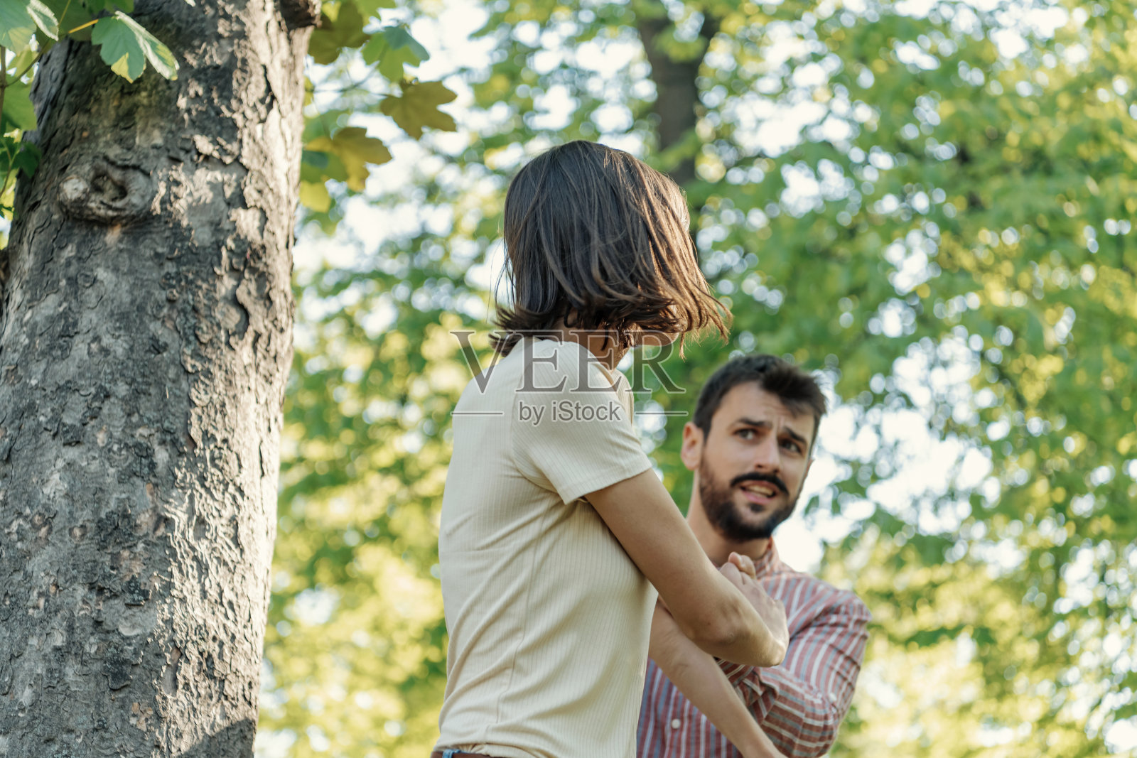一对年轻夫妇在公园里吵架。年轻夫妇有一个夏天的一天争吵。站在公园外面，困惑的年轻女子打着手势，看着她的男朋友照片摄影图片