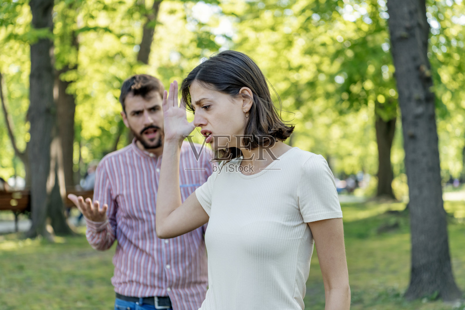 一对年轻夫妇在公园里吵架。年轻夫妇有一个夏天的一天争吵。站在公园外面，困惑的年轻女子打着手势，看着她的男朋友照片摄影图片