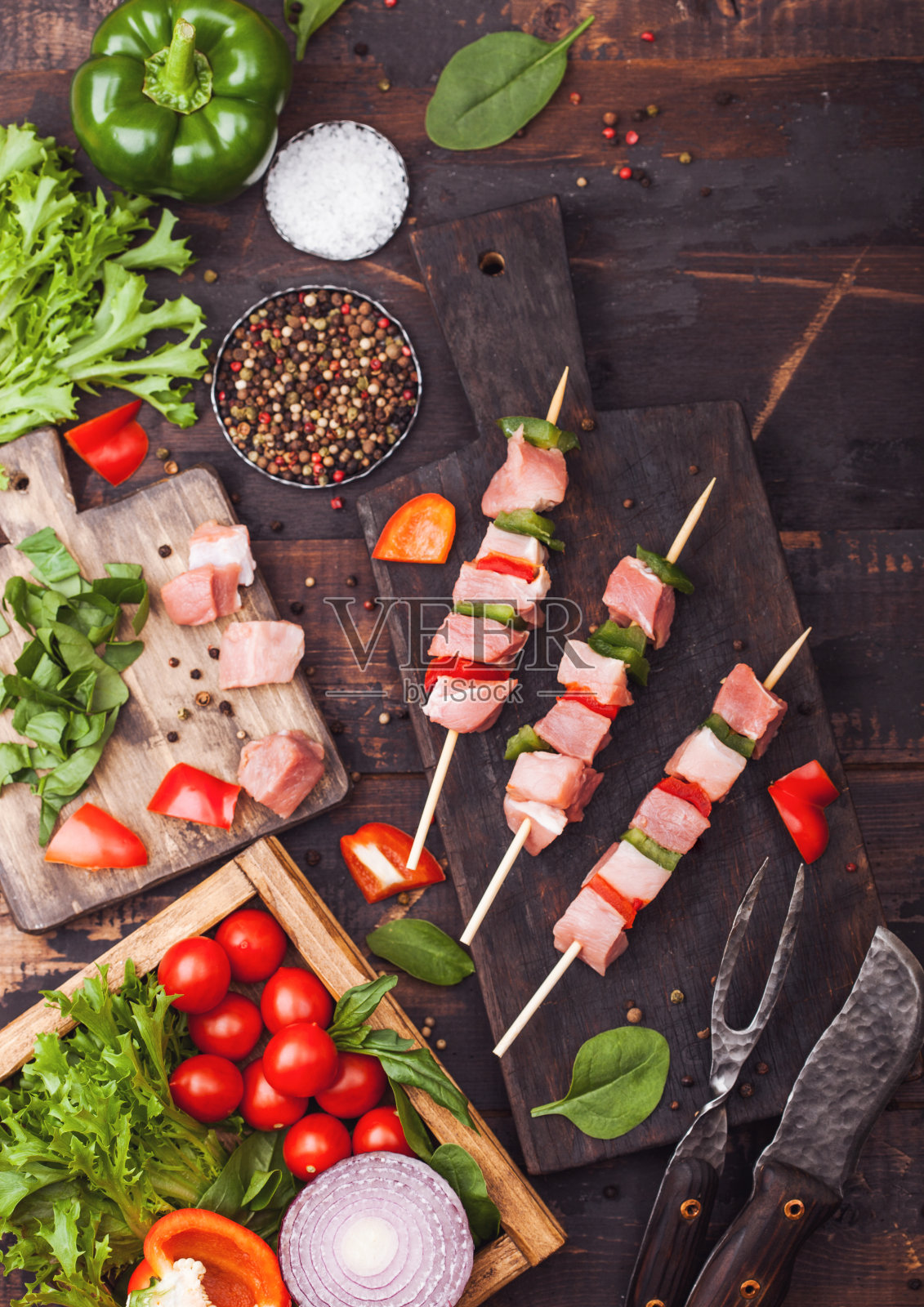 切菜板上放着红辣椒的生猪肉串，木背景上放着新鲜蔬菜，用刀叉。盐、胡椒粉、生菜、辣椒粉和圣女果。俯视图照片摄影图片