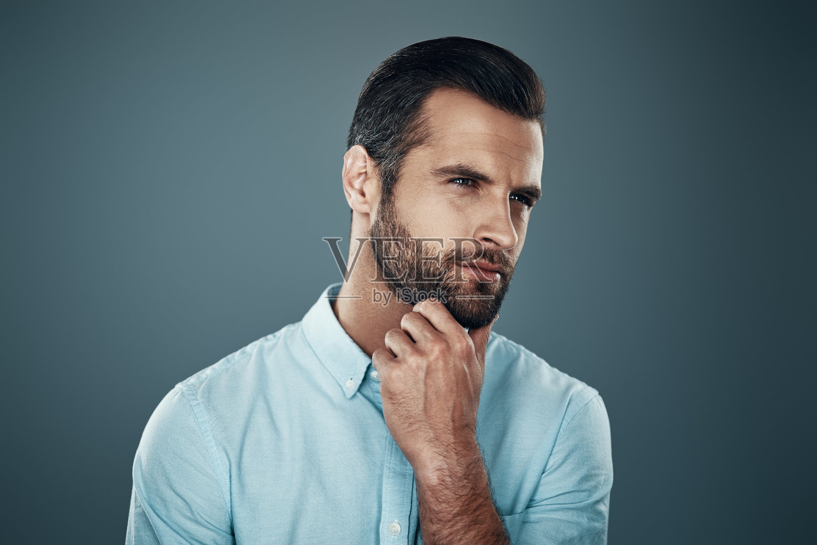 展望未来。英俊的年轻人望着别处，手放在下巴上，站在灰色的背景下照片摄影图片