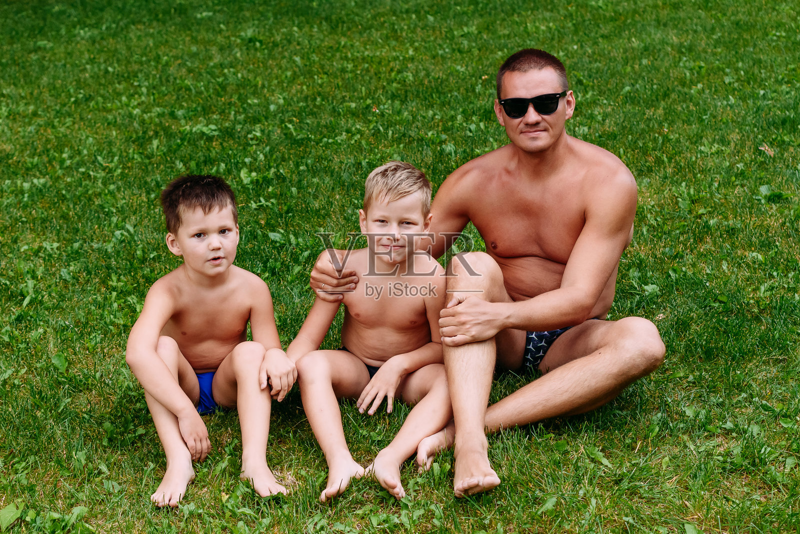两个男孩和一个穿泳裤的男人在夏天坐在草坪上。快乐的人,照片摄影图片