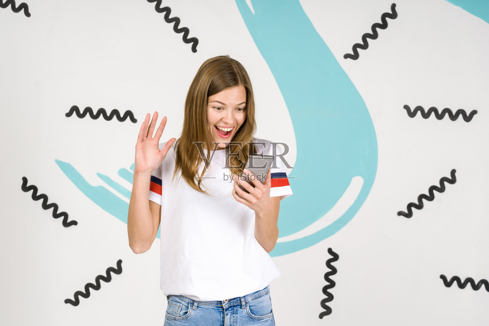 年轻美丽的女子站在白色的艺术背景上，手里拿着智能手机，在镜头前挥手问好照片摄影图片