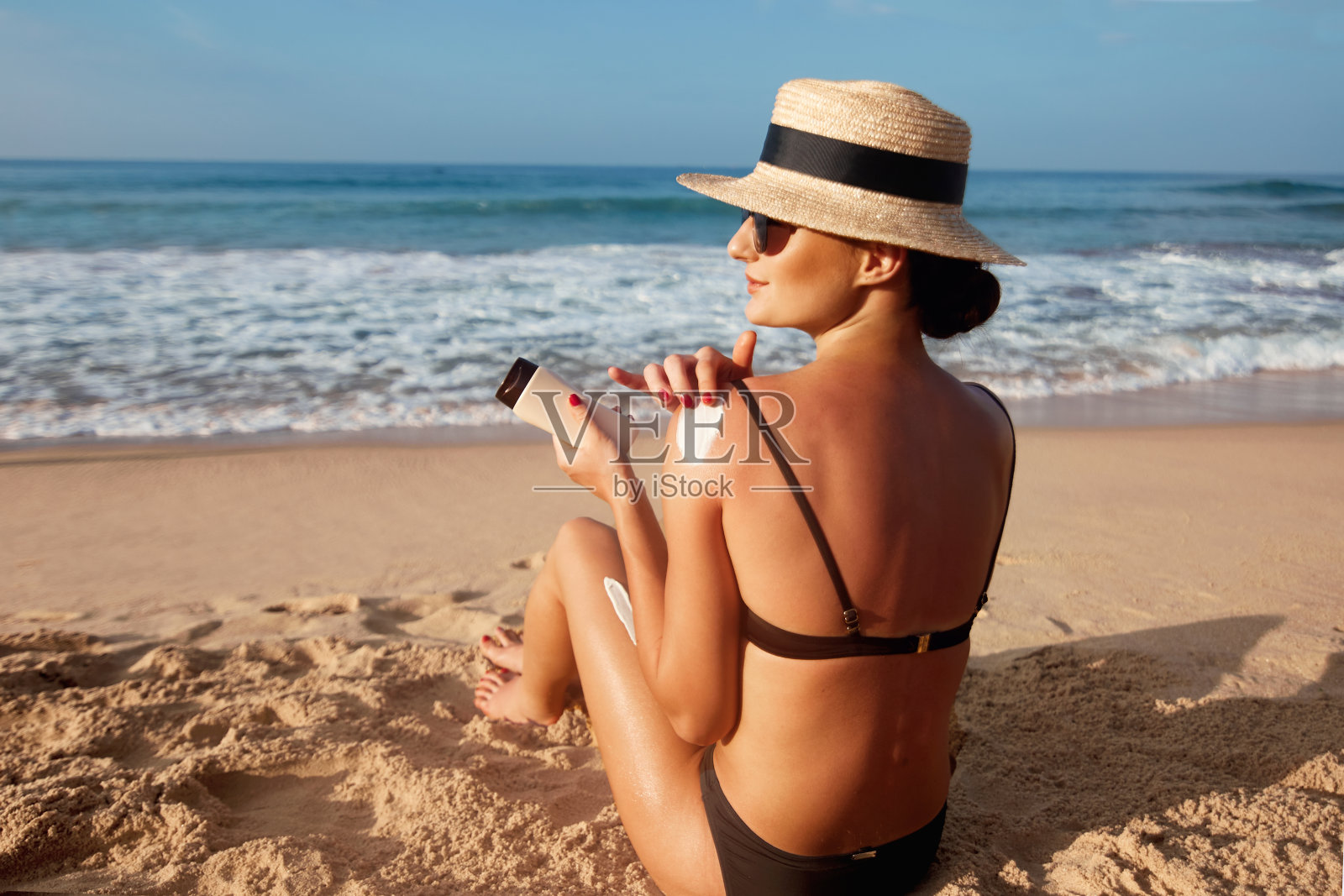 美女在晒黑的肩膀上涂防晒霜。护肤品。身体防晒防晒霜。比基尼帽子妇女使用保湿防晒乳液背部照片摄影图片