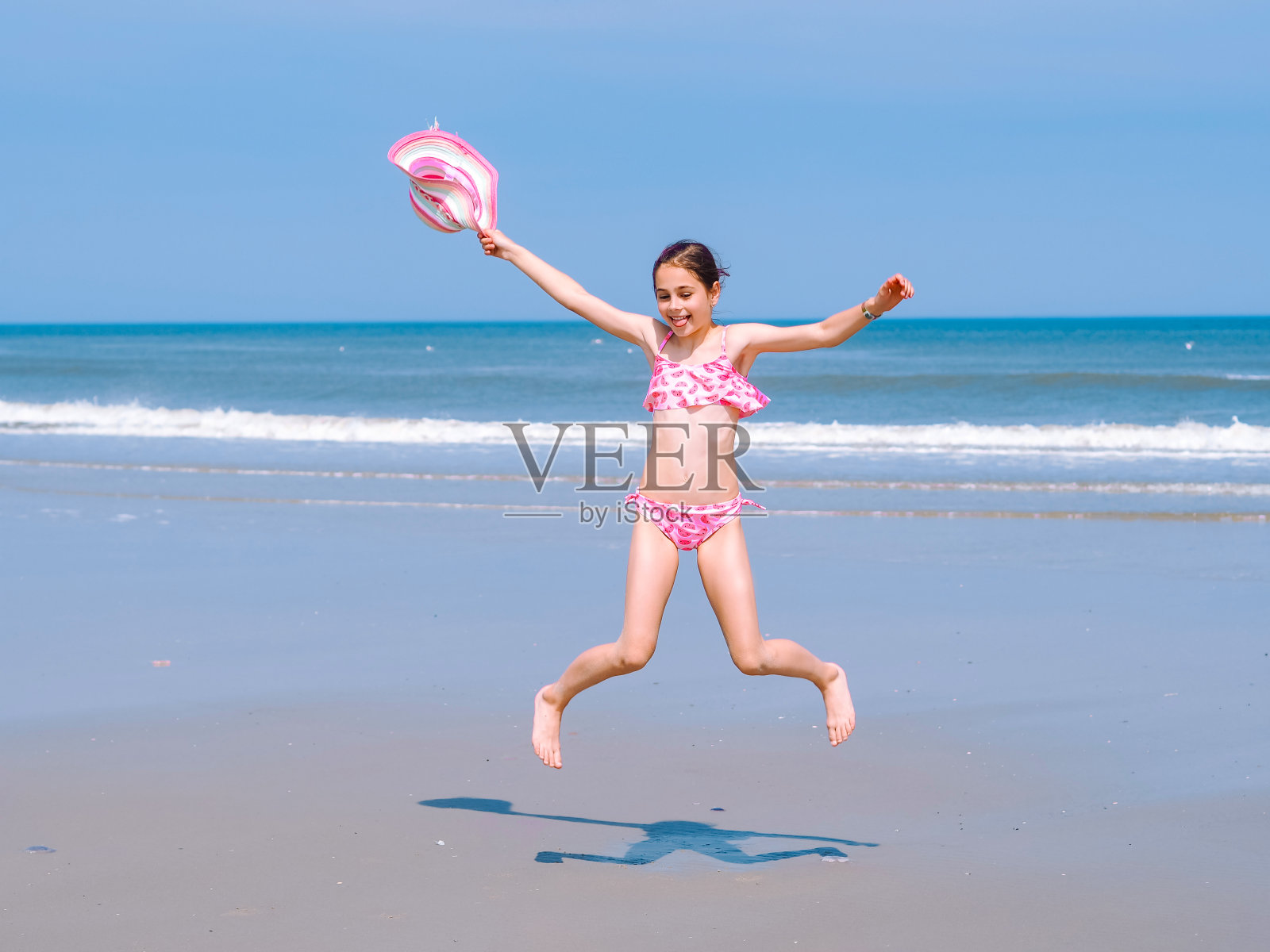年轻快乐的十几岁的女孩有乐趣的热带海滩和跳在粉红色的泳衣和条纹帽子到海边的空中照片摄影图片