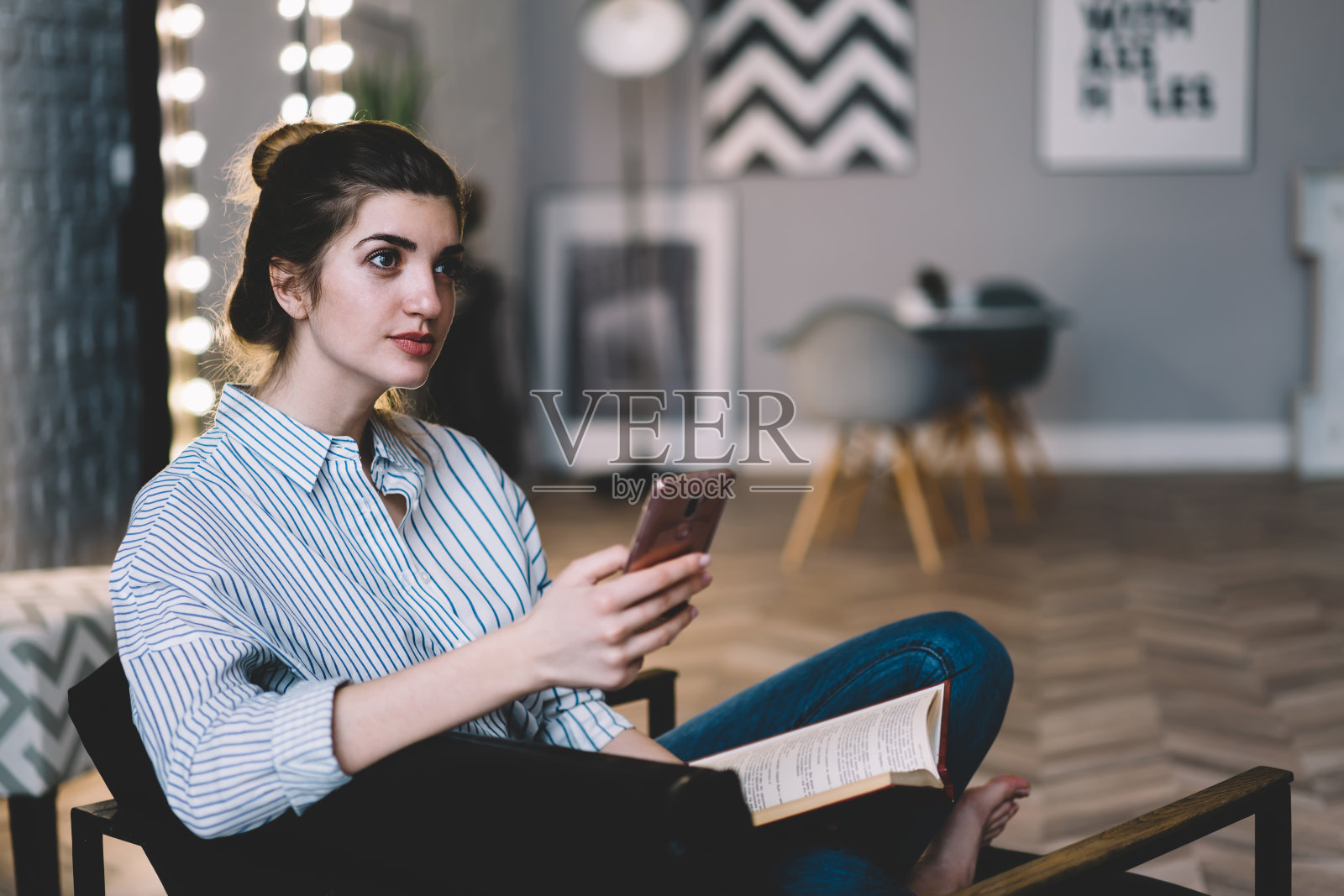 美丽聪明的年轻女子的书籍爱好者沉思在智能手机收到通知。迷人的潮女手拿手机，在时尚的室内享受休闲时光照片摄影图片