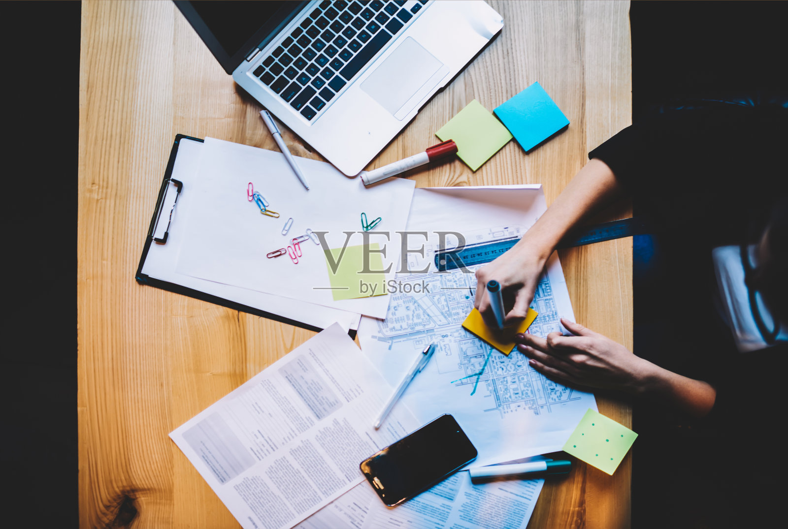 木质桌面俯视图，带有建筑草图和现代笔记本电脑设备键盘。女人的手画在彩色贴纸上的标记坐在桌子上与必要的办公设备照片摄影图片