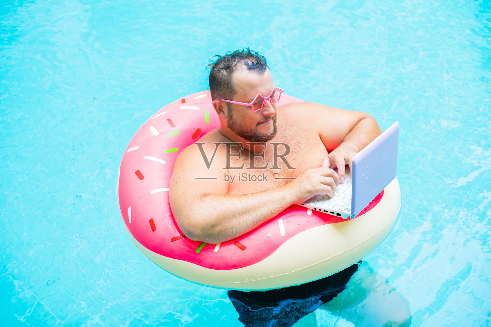 一个戴着粉色眼镜的搞笑胖男人在游泳池里的一个充气圈里工作，在笔记本电脑上画一个女孩。照片摄影图片