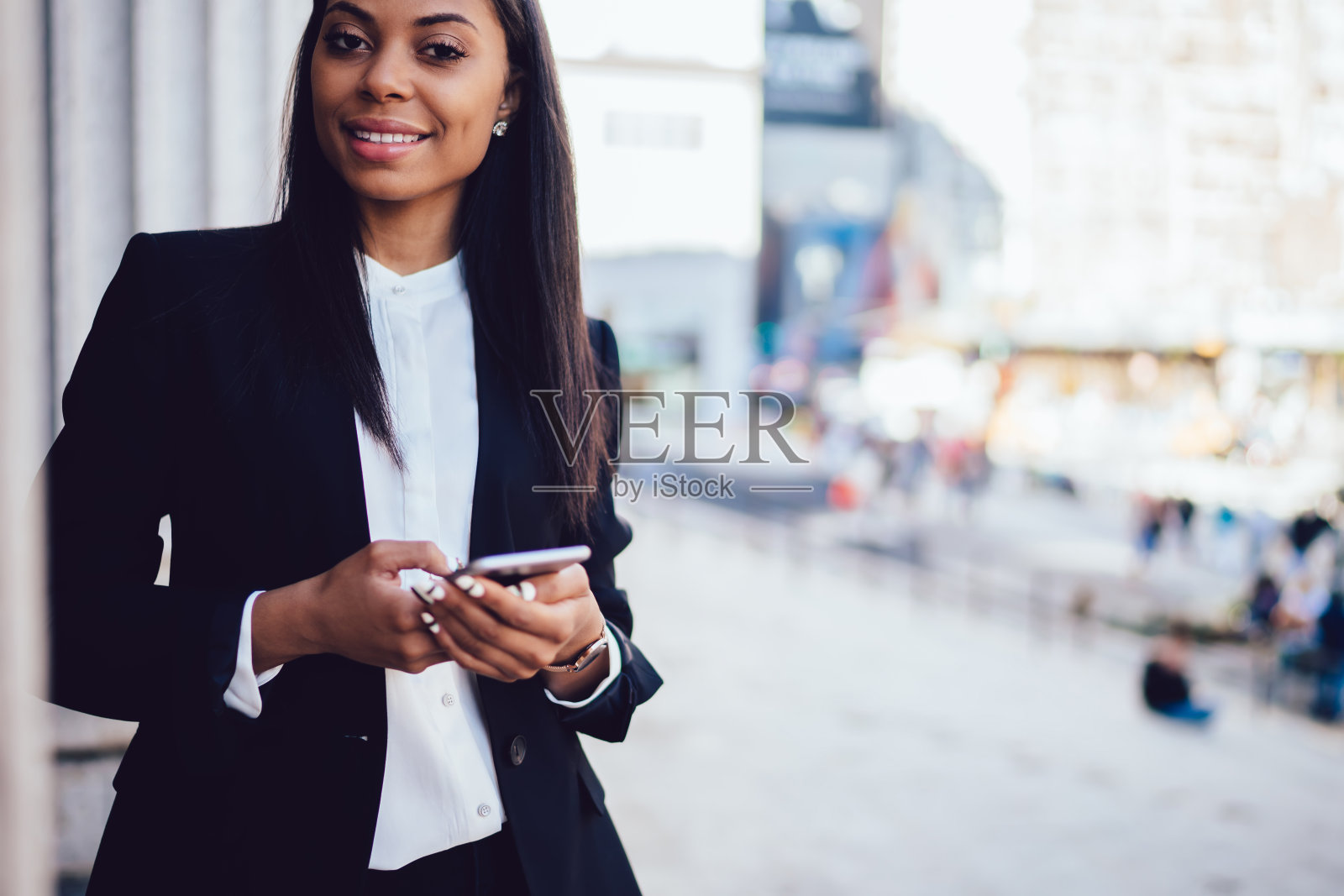 这是一位成功的非裔美国女企业家的肖像，她穿着时尚的正装，拿着智能手机，对着镜头微笑，站在街上。商人使用手机照片摄影图片