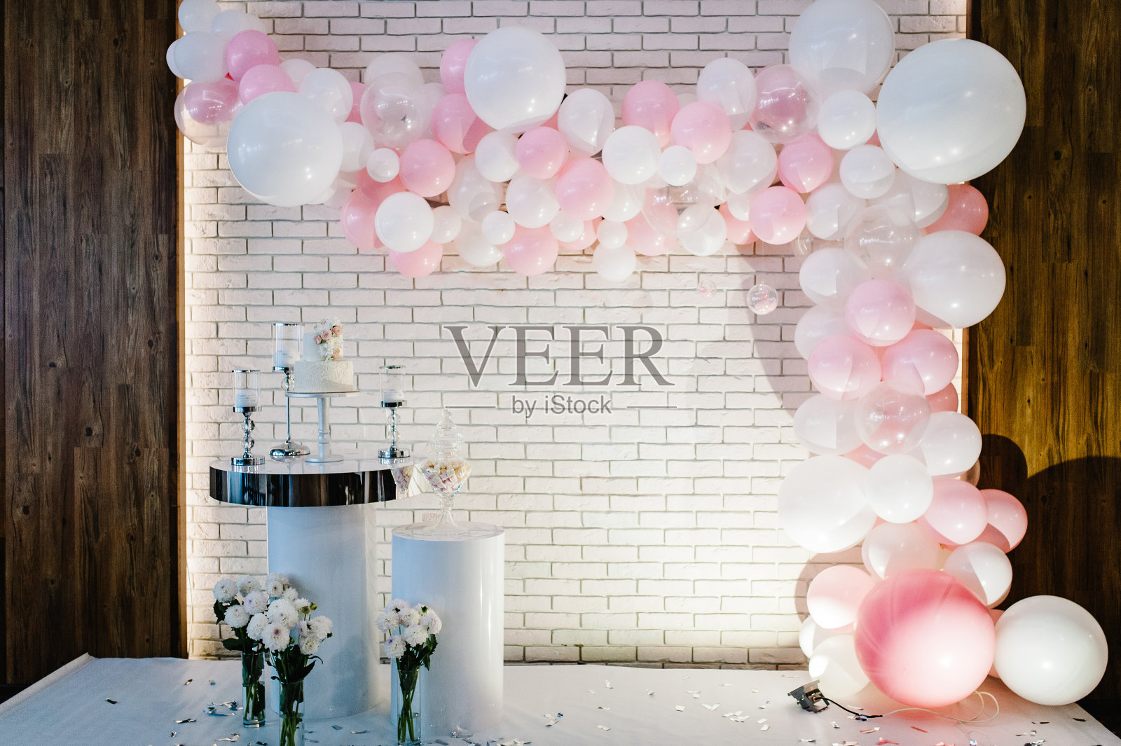 照片墙，婚礼装饰空间或地方从白色和粉色的气球和白色的砖墙附近的桌子上有一个婚礼蛋糕，蜡烛和鲜花。照片摄影图片
