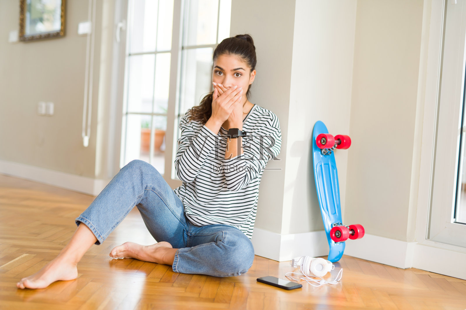 年轻女人坐在地板上玩滑板，戴着耳机，因犯错而捂住嘴巴，秘密概念照片摄影图片