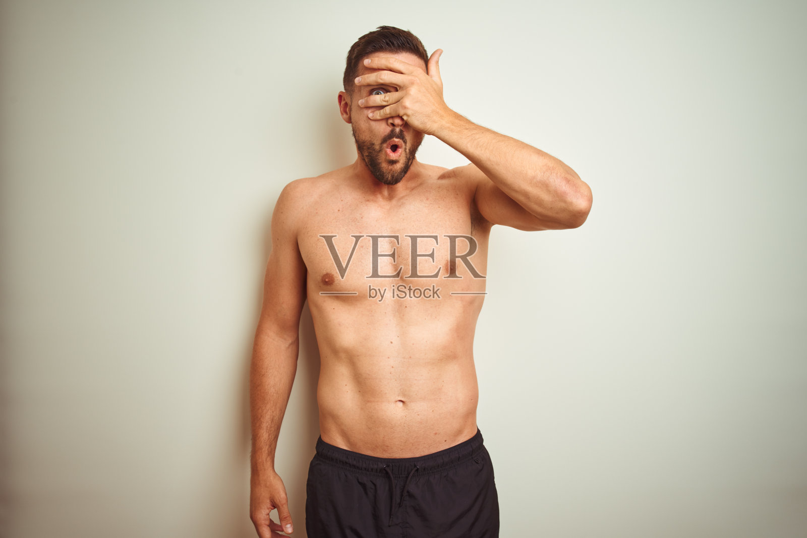 年轻英俊的赤膊男子在孤立的背景中窥视，用手捂着脸和眼睛，透过手指看着尴尬的表情。照片摄影图片