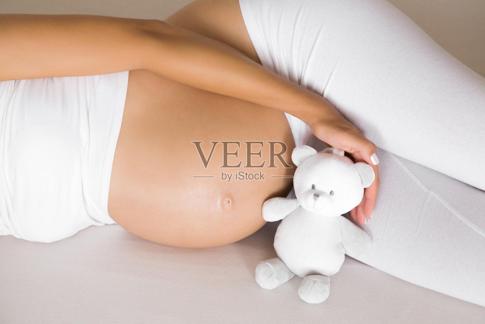 女人赤裸着肚子躺在床上。手牵着小微笑的白色泰迪熊为未来的宝宝。怀孕35周的时候，充满爱意的时刻。孩子的期望。前视图。特写镜头。照片摄影图片