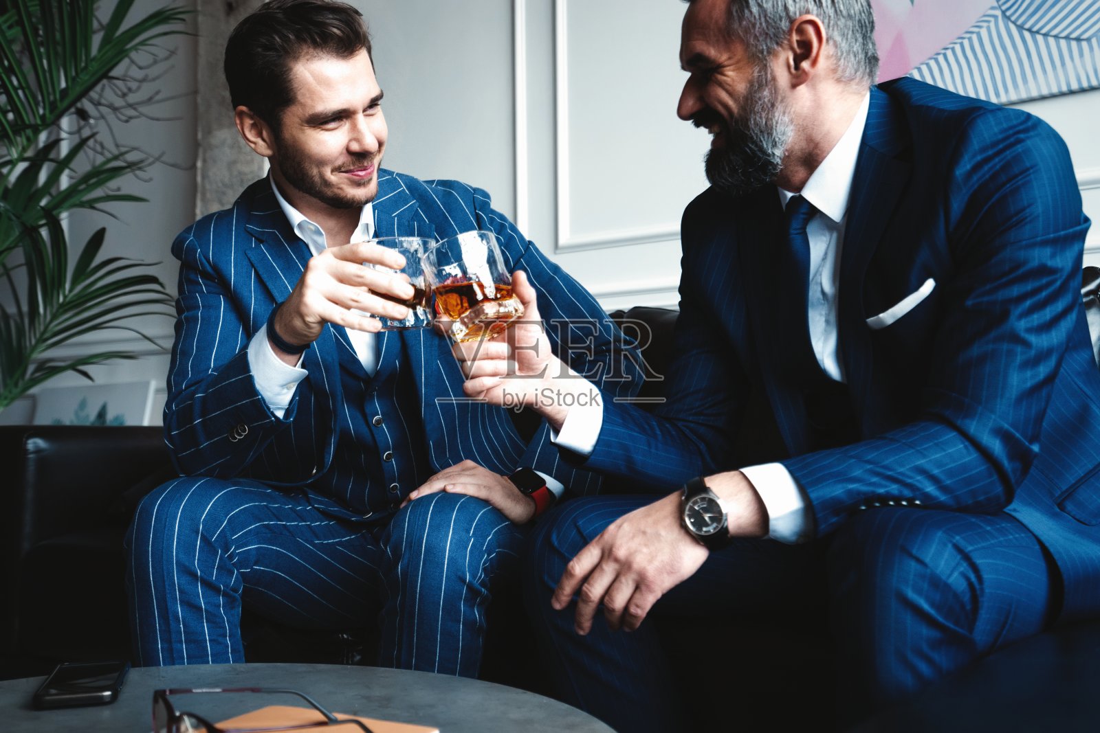 放松。在室内休息时，两个年轻英俊的男人穿着西装，拿着眼镜，看着对方。照片摄影图片