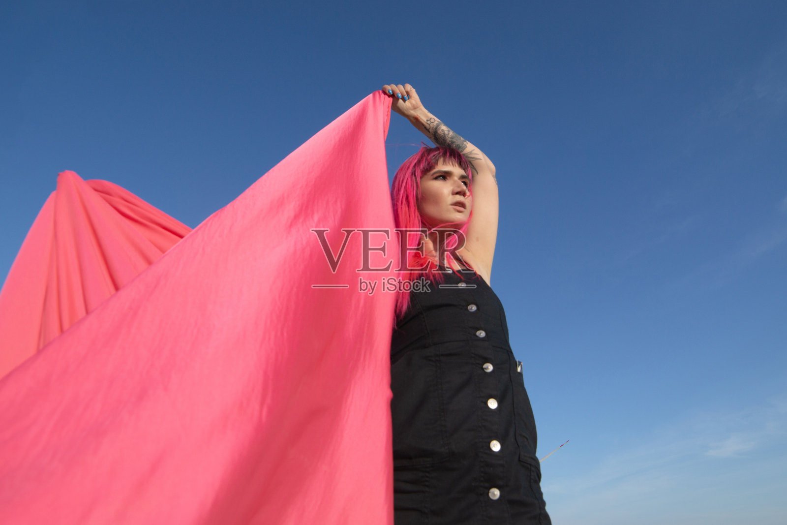 粉红色头发的年轻女子用粉红色的布片和风力发电机在田地附近跳舞。风车在日落。时尚的肖像照片摄影图片