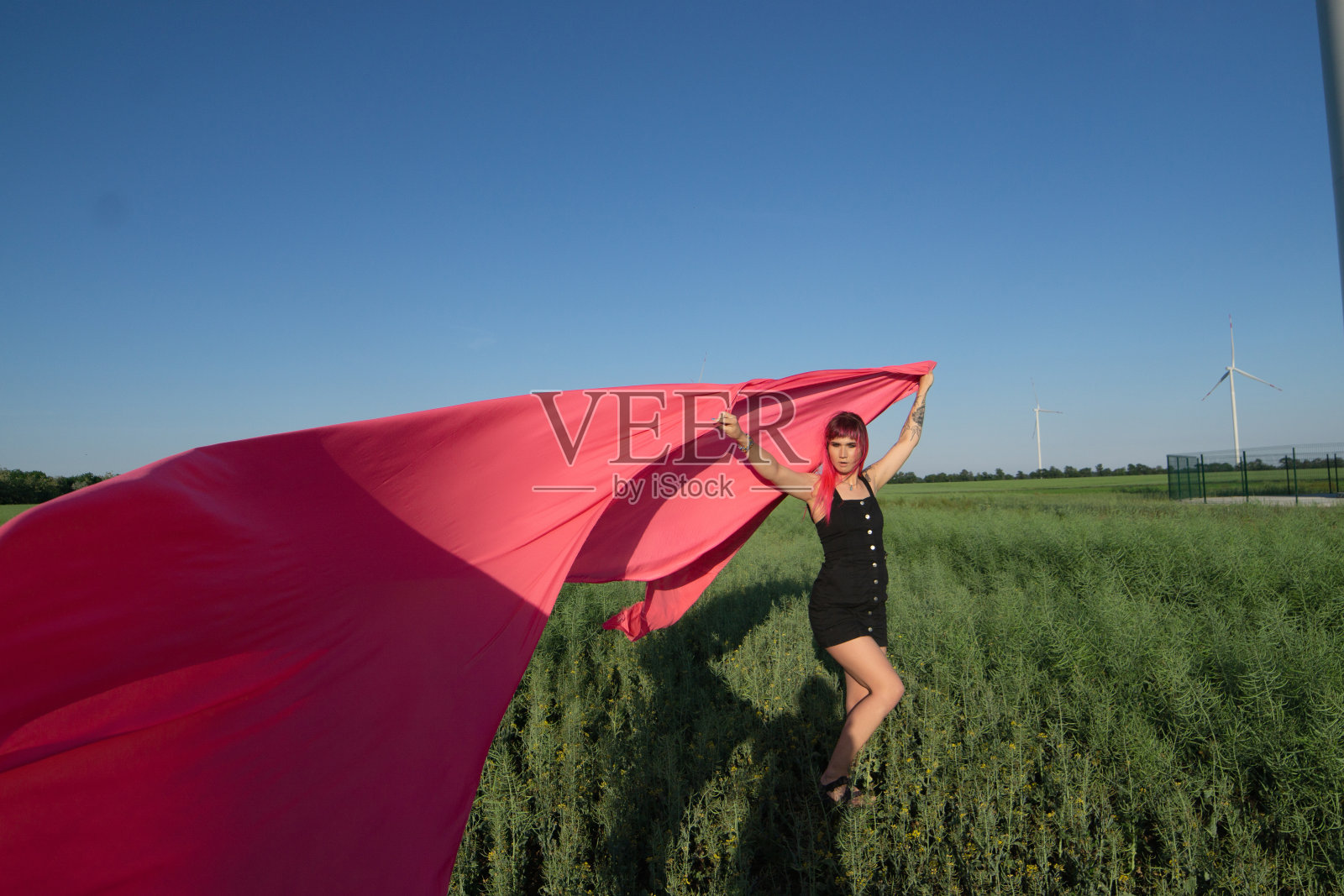 粉红色头发的年轻女子用粉红色的布片和风力发电机在田地附近跳舞。风车在日落。时尚的肖像照片摄影图片