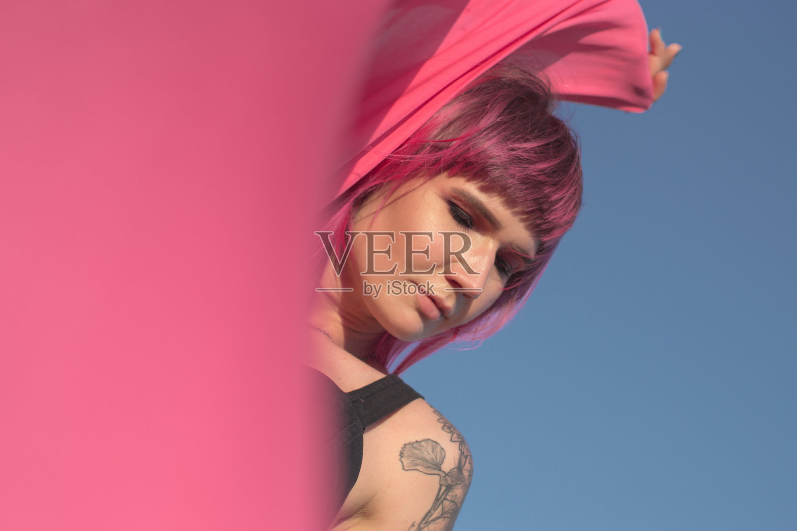 粉红色头发的年轻女子用粉红色的布片和风力发电机在田地附近跳舞。风车在日落。时尚的肖像照片摄影图片