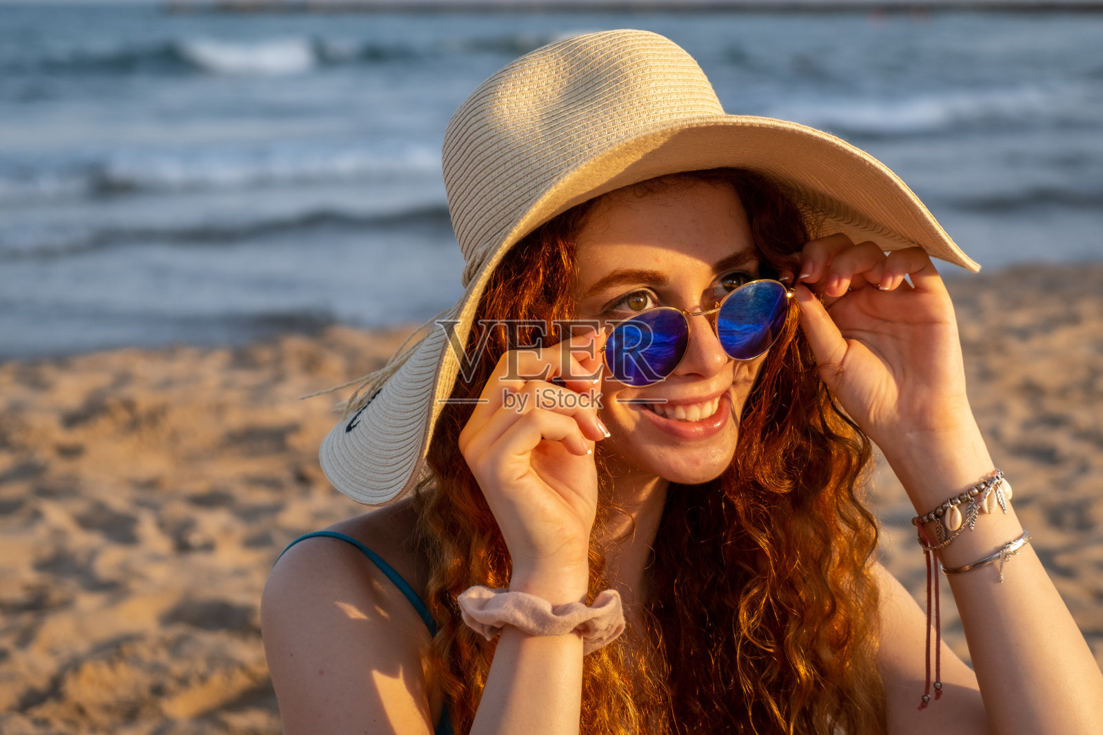 年轻的红发女孩戴着帽子和太阳镜在地中海的海滩上。照片摄影图片