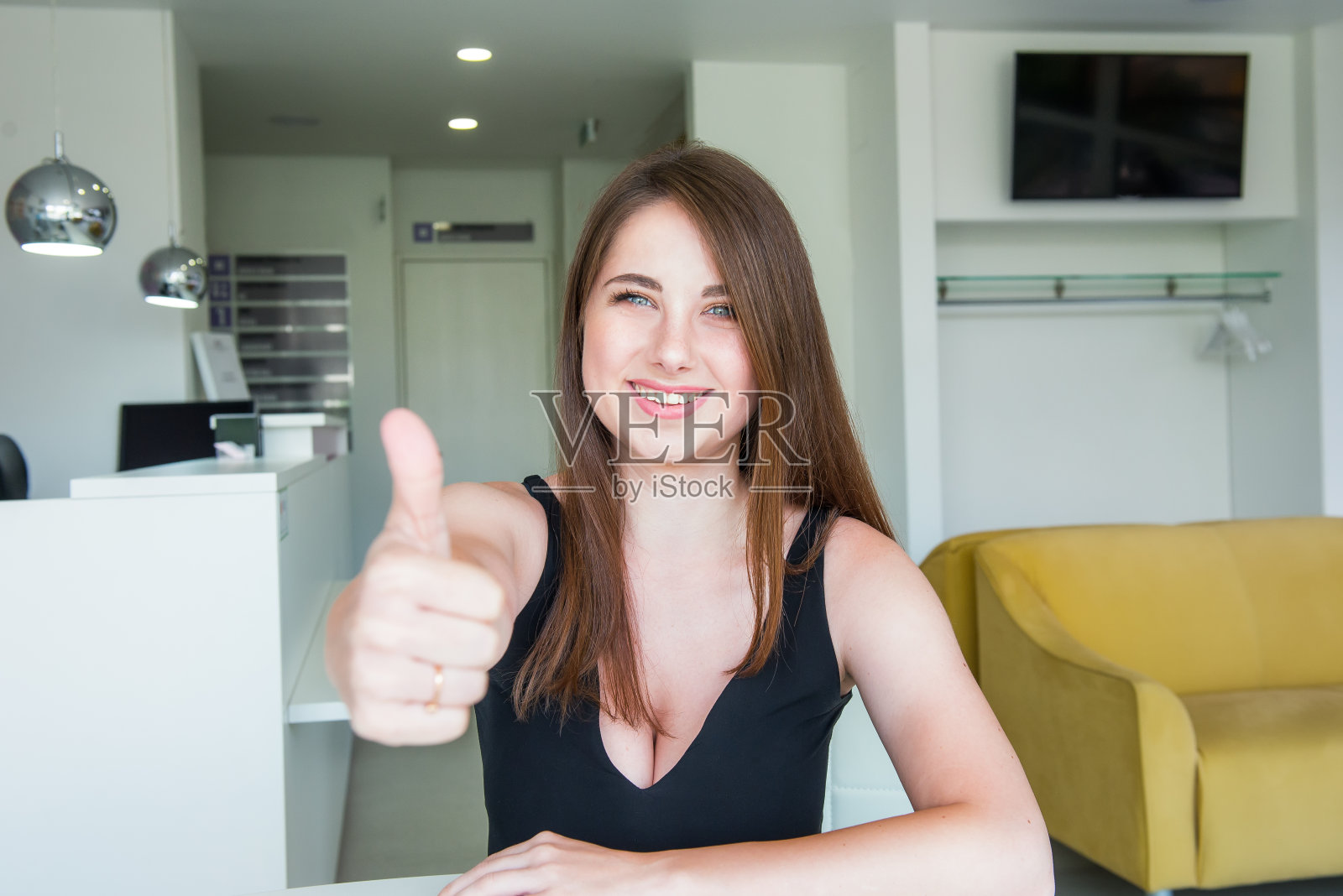 近距离的肖像快乐微笑的年轻女子与深胸和拇指向上的手坐在办公桌后面在办公室大厅。选择性聚焦，复制空间。照片摄影图片