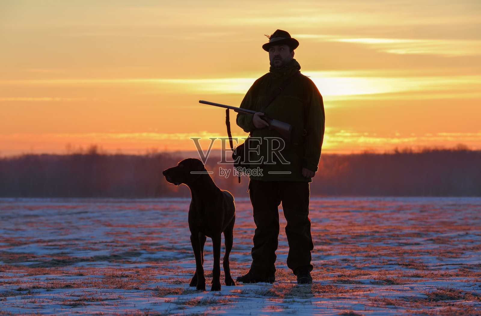 一个戴着帽子拿着枪的猎人和一只德国短毛犬站在一片覆盖着雪的草地上。雄伟的冬季景色。神奇的日出。照片摄影图片