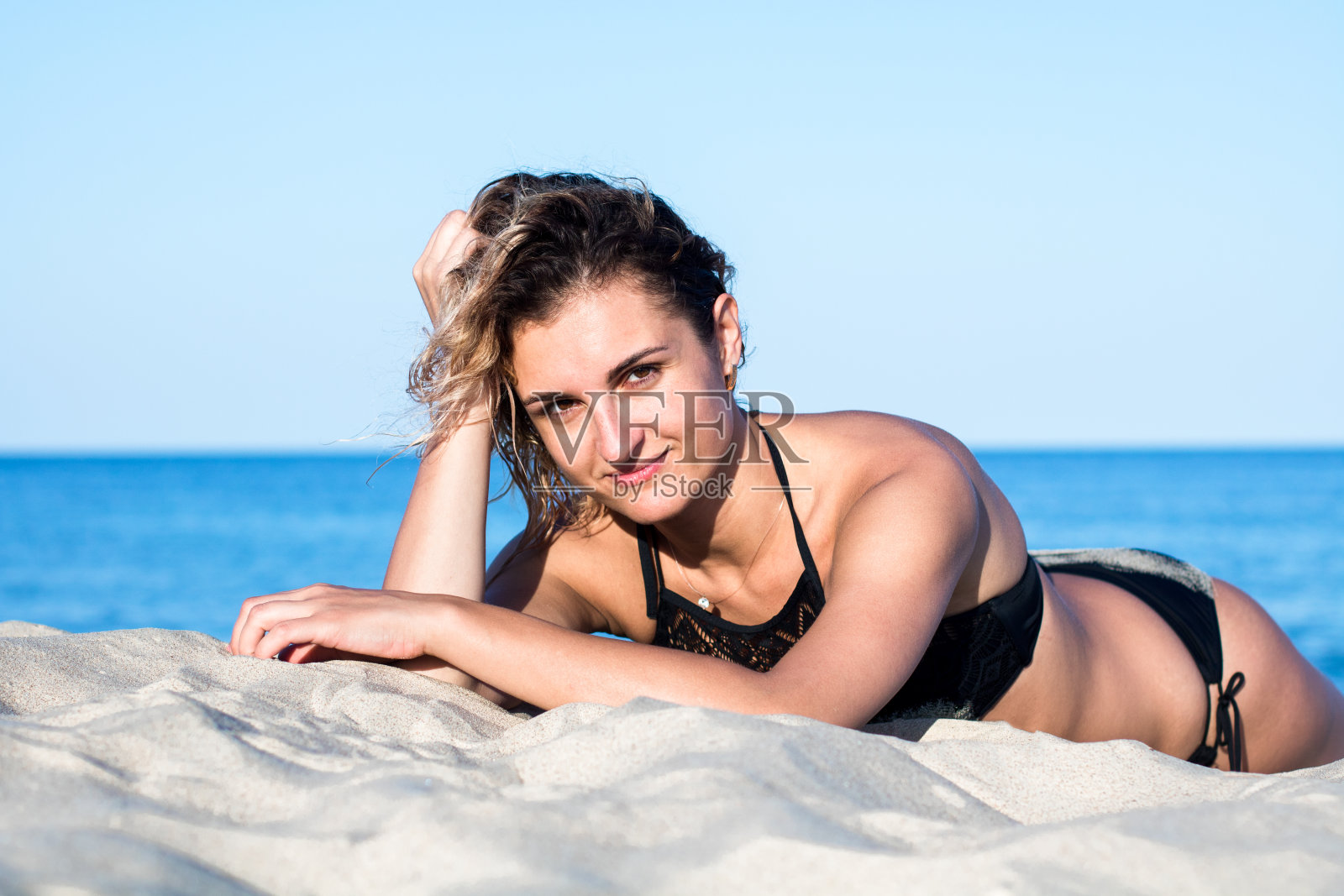 古铜色的年轻迷人的女人卷发穿着黑色泳衣在沙滩上，躺在沙滩上照片摄影图片