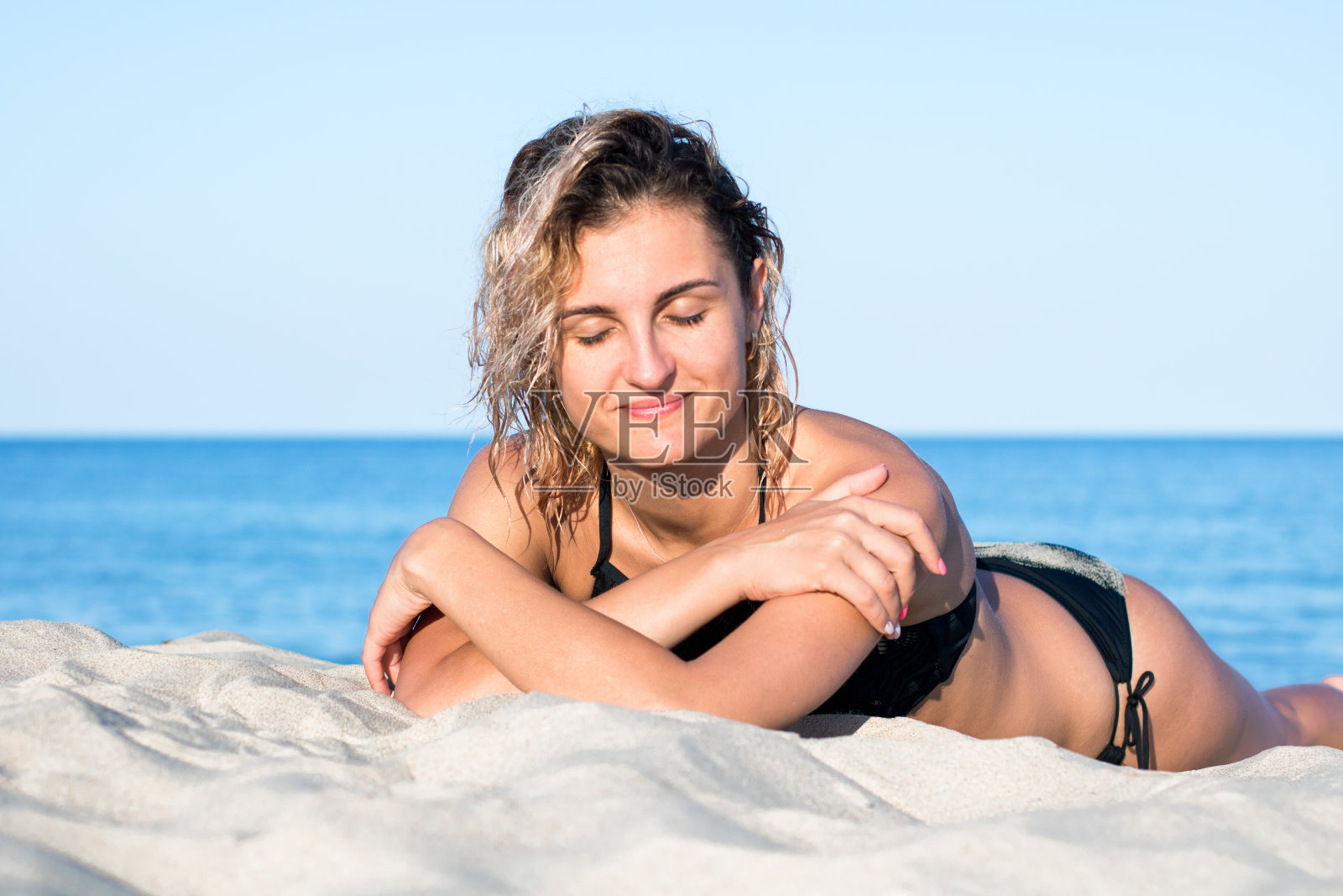 古铜色的年轻迷人的女人卷发穿着黑色泳衣在沙滩上，躺在沙滩上照片摄影图片