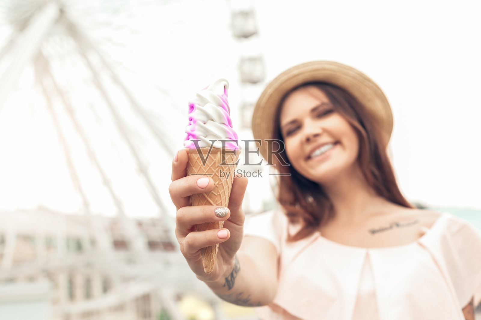 户外休闲。戴着帽子的女孩走在摩天轮附近的街道上，带着冰淇淋蛋卷迷失了模糊的背景，微笑着照片摄影图片