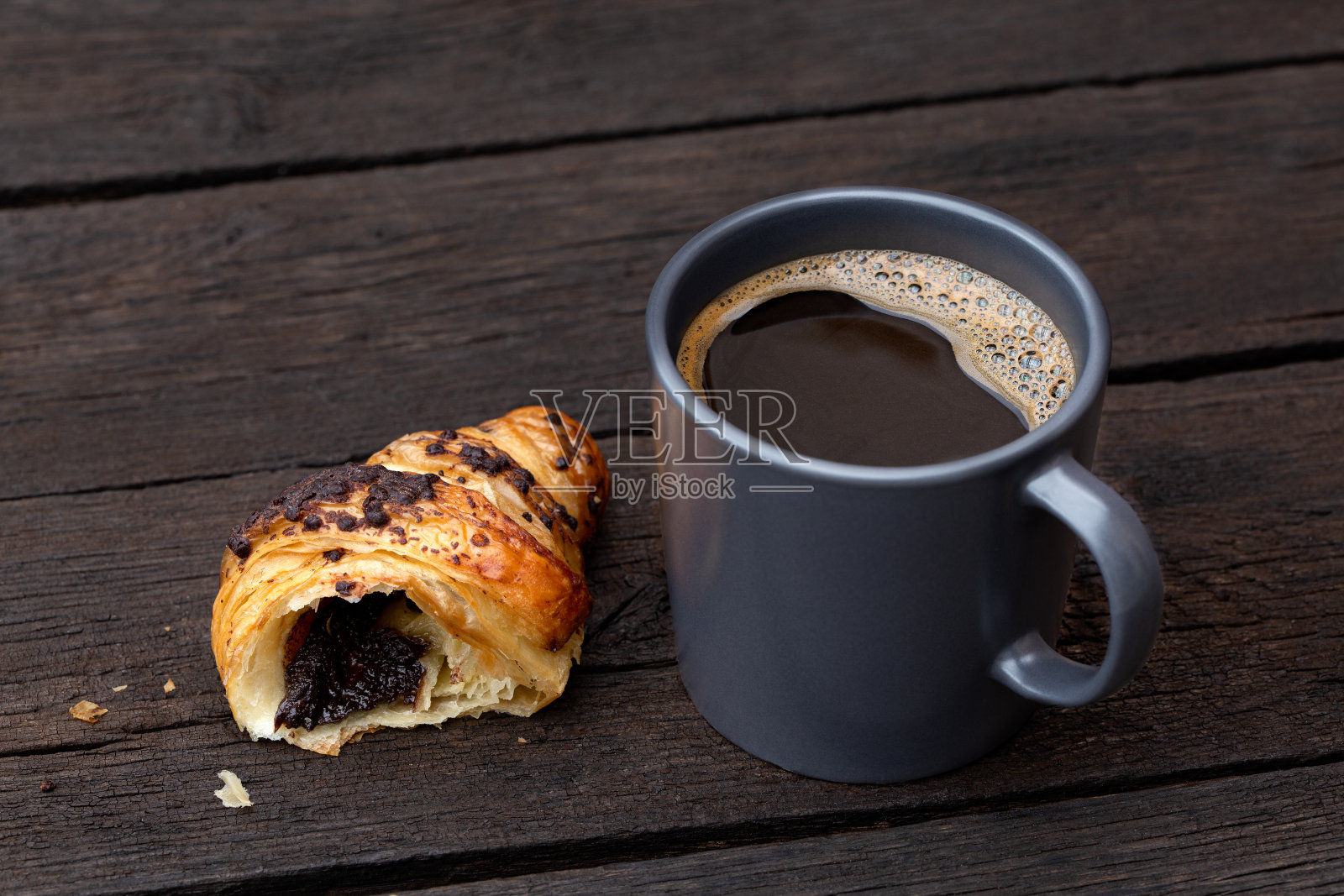 蓝灰色的陶瓷杯里装着黑咖啡，半块巧克力羊角面包单独放在质朴的深褐色木桌上。照片摄影图片