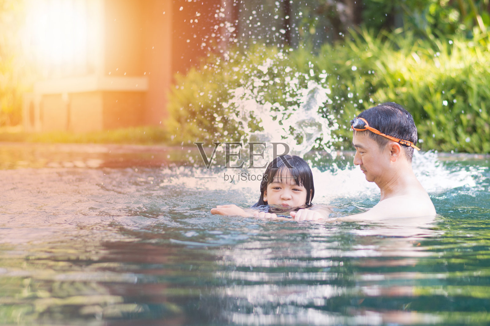 女儿挽着爸爸的胳膊练习游泳。爸爸在自然度假酒店教小女孩游泳。教练让孩子在游泳池里踢腿泼水。照片摄影图片