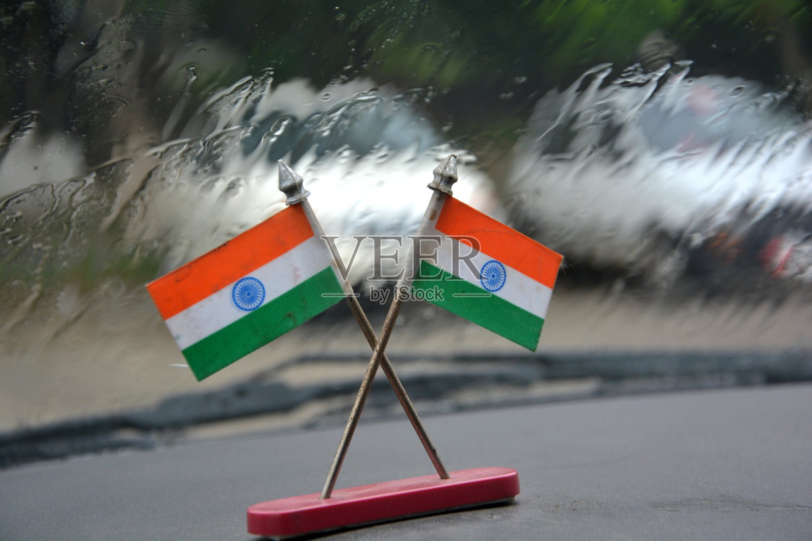 两个微型印度国旗的特写，由纸制成的小金属支架交叉在一个汽车仪表板的顶部，背景水从挡风玻璃上落下，选择性聚焦照片摄影图片