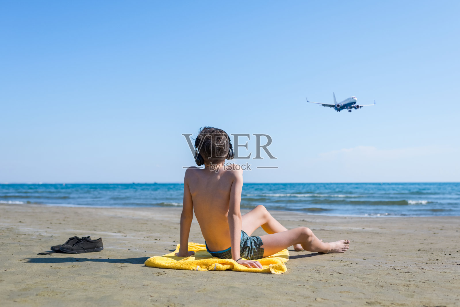 男孩坐在黄色的毛巾上戴着耳机，用手指在沙滩上展示着飞机降落的海面和天空的背景。旅游的概念照片摄影图片