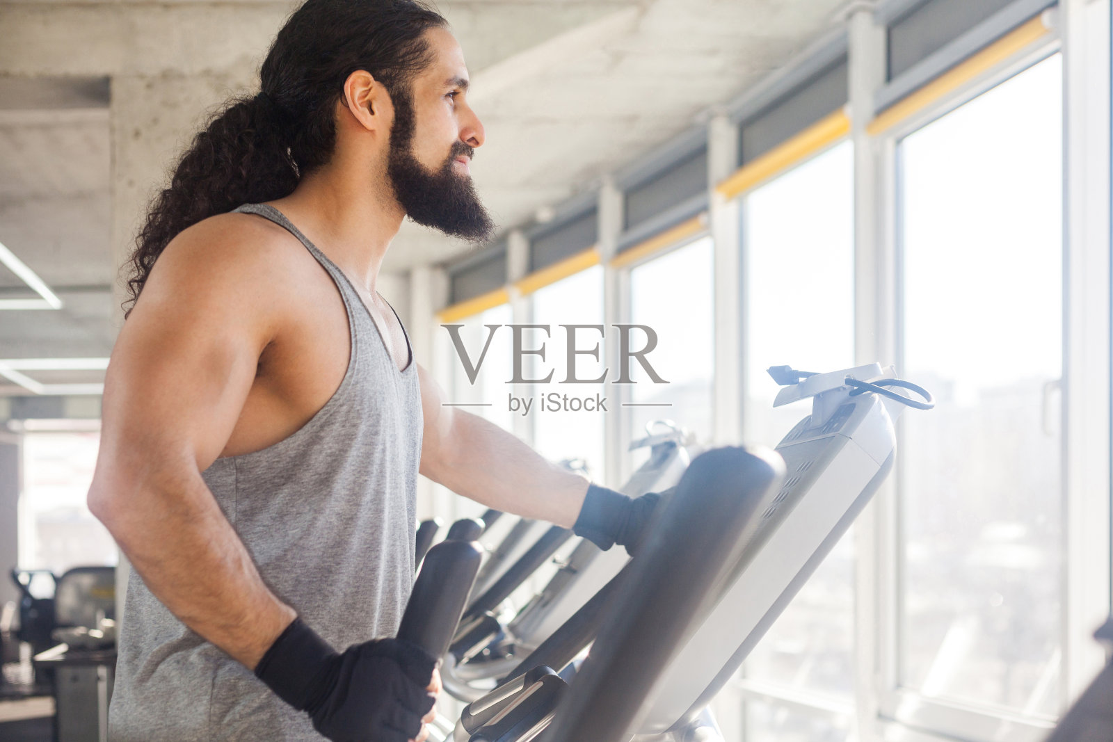 年轻成年运动男子长卷发独自训练的近距离侧面 портрет。英俊的男人在椭圆机上在健身房锻炼，看着窗户。照片摄影图片